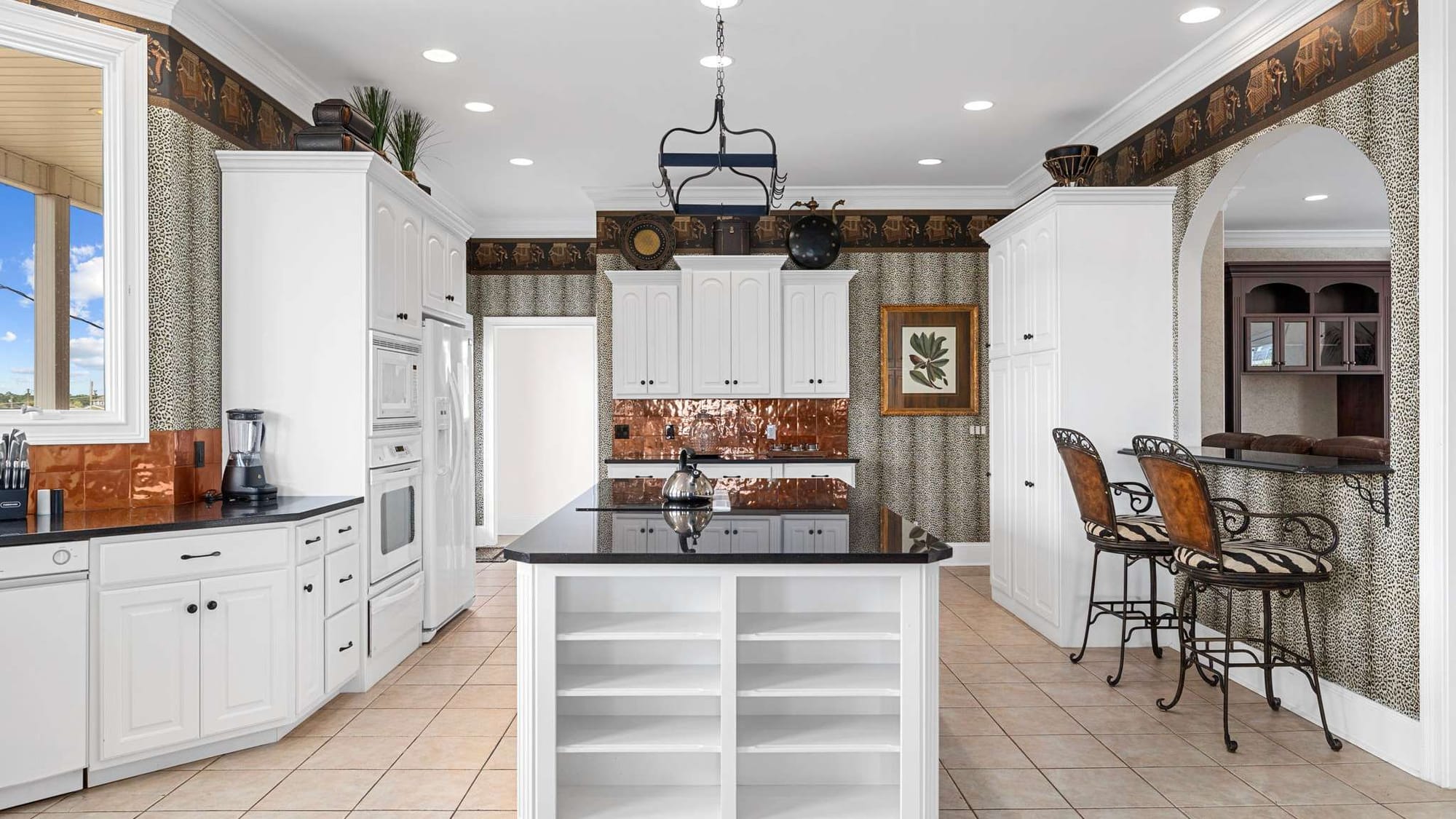 A spacious kitchen with white cabinetry, black countertops, copper backsplash accents, and a central island with open shelving, featuring animal-print wallpaper and high-back bar stools.