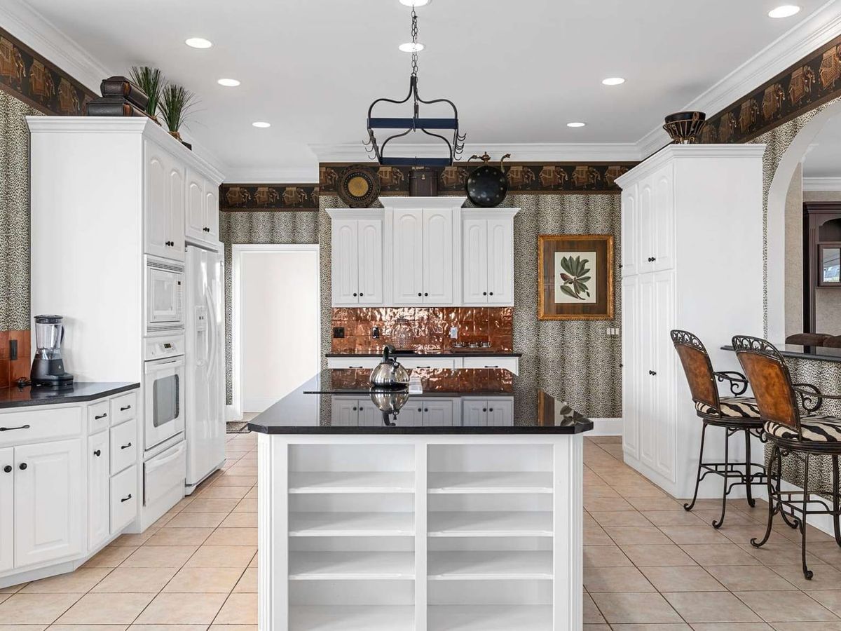 A spacious kitchen with white cabinetry, black countertops, copper backsplash accents, and a central island with open shelving, featuring animal-print wallpaper and high-back bar stools.