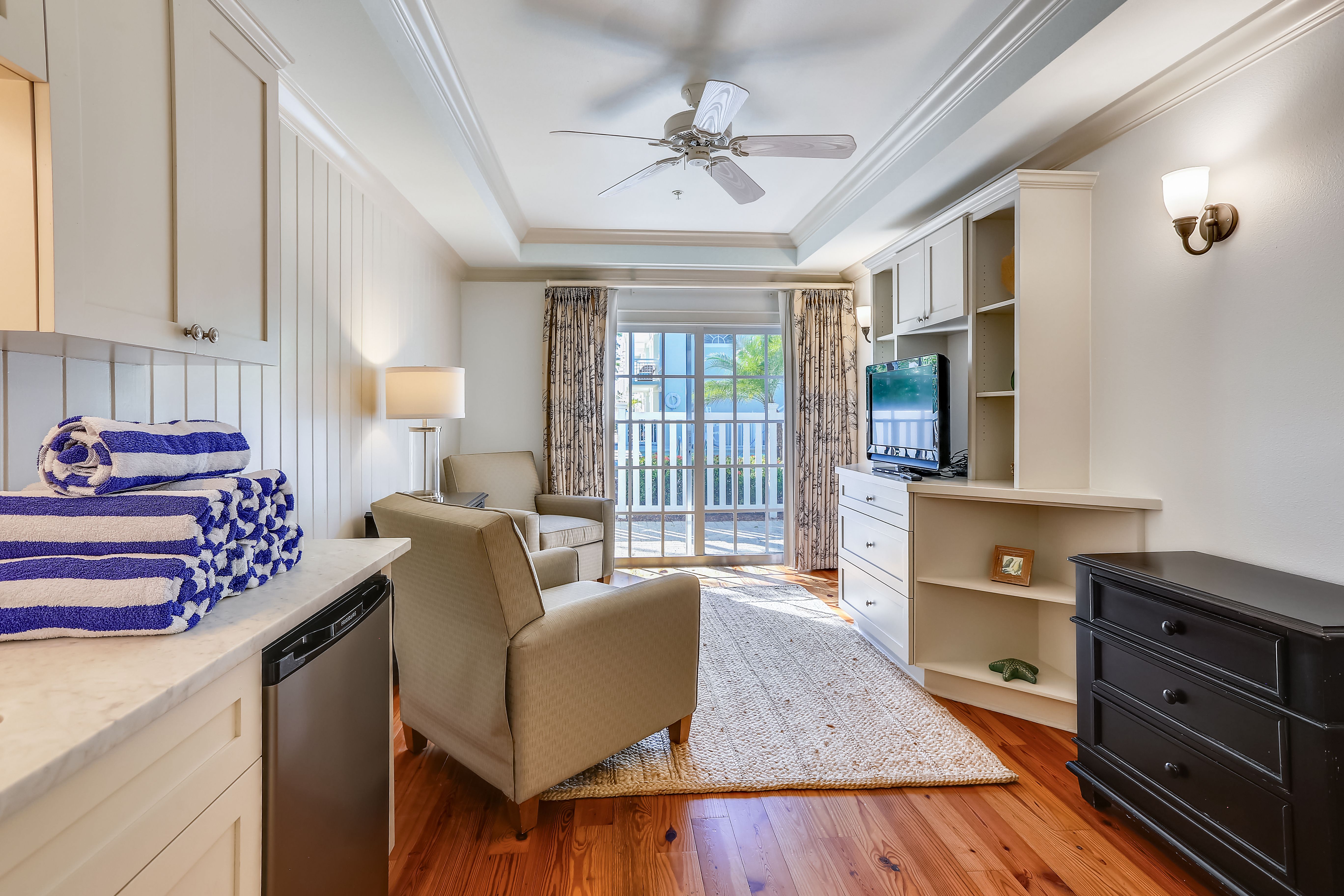Cozy and sunlit coastal den featuring two armchairs, a flat-screen TV, and French doors leading to a private patio. A stack of striped pool towels adds a splash of beachy charm to the light, airy decor and warm wood floors.