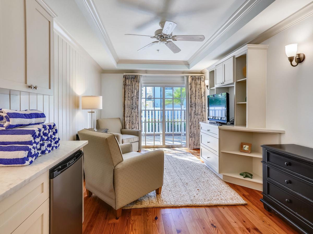 Cozy and sunlit coastal den featuring two armchairs, a flat-screen TV, and French doors leading to a private patio. A stack of striped pool towels adds a splash of beachy charm to the light, airy decor and warm wood floors.
