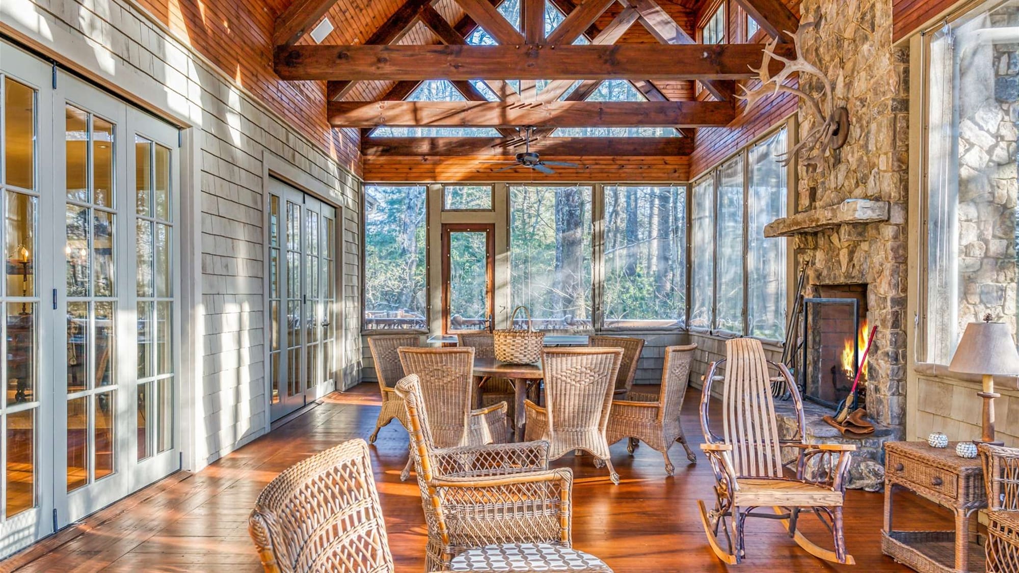 A bright sunroom with wooden beams and a stone fireplace offers cozy seating surrounded by forest views. Natural light pours in through large windows, making it a warm and inviting mountain retreat. It’s a peaceful space to gather, relax, and enjoy cabin life in the woods.