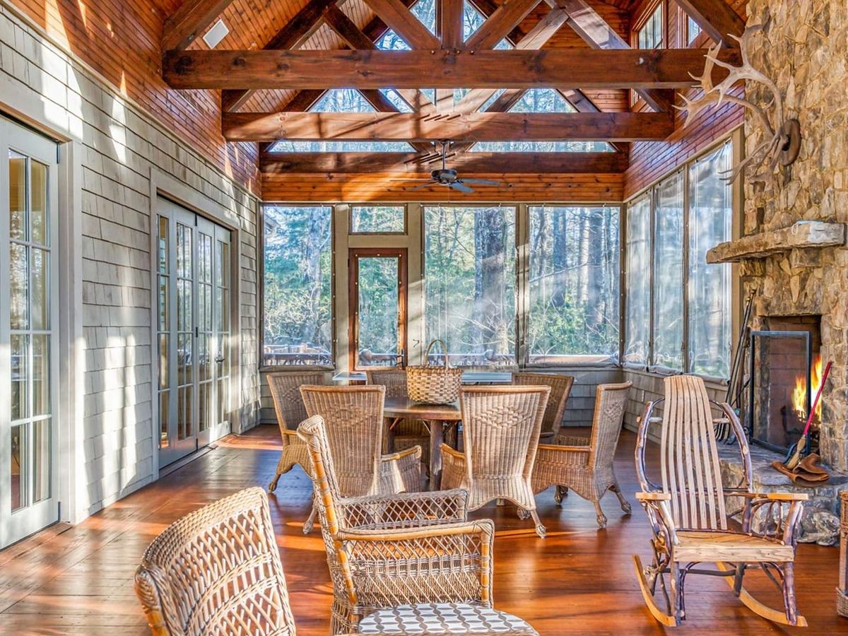 A bright sunroom with wooden beams and a stone fireplace offers cozy seating surrounded by forest views. Natural light pours in through large windows, making it a warm and inviting mountain retreat. It’s a peaceful space to gather, relax, and enjoy cabin life in the woods.
