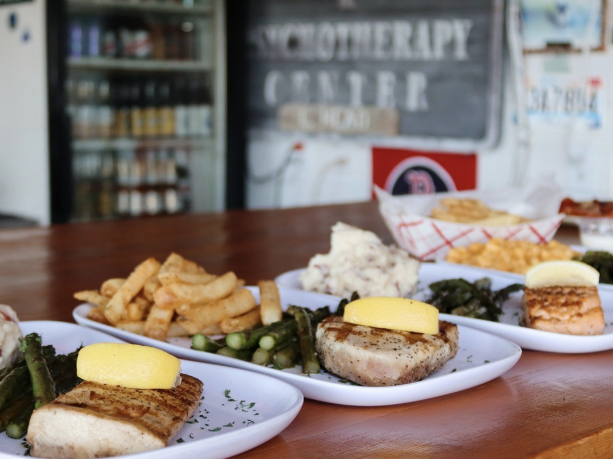 A row of seafood plates featuring grilled fish topped with lemon wedges, served with sides like asparagus, fries, mashed potatoes, and greens on a wooden counter in a casual restaurant setting.