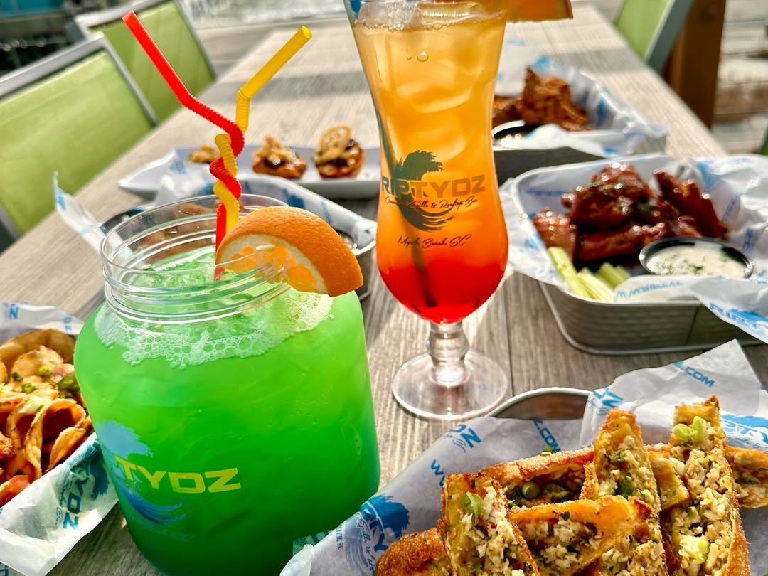Colorful cocktails paired with fried seafood egg rolls and dipping sauce, with a beautiful ocean view in the background. RipTydz combines lively rooftop vibes with classic beach eats and drinks.