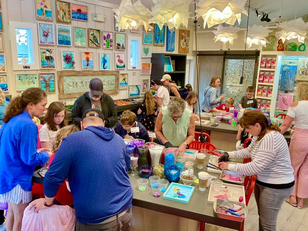 Families and kids gather around colorful workstations inside The Shard Shop, creating handmade glass art. The bright space is decorated with vibrant ocean-inspired artwork, offering a fun and hands-on creative experience.