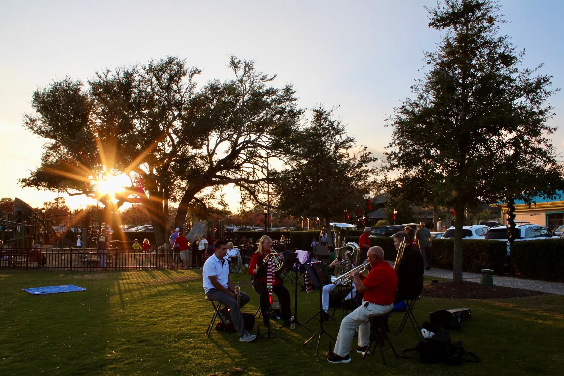 A small brass ensemble performs at sunset on the lawn at Barefoot Landing in Myrtle Beach, with golden rays of sunlight streaming through live oak trees and holiday lights decorating the outdoor shopping village in the background