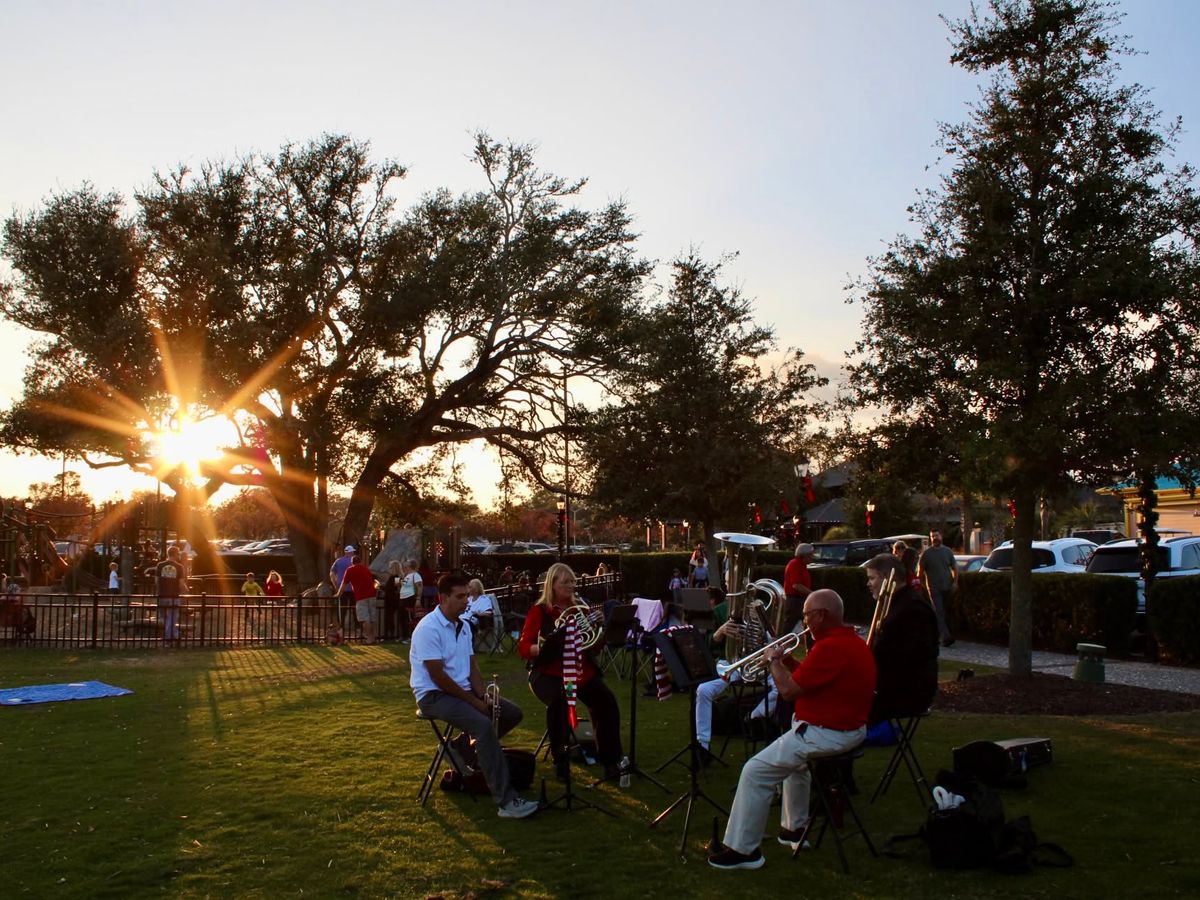 A small brass ensemble performs at sunset on the lawn at Barefoot Landing in Myrtle Beach, with golden rays of sunlight streaming through live oak trees and holiday lights decorating the outdoor shopping village in the background
