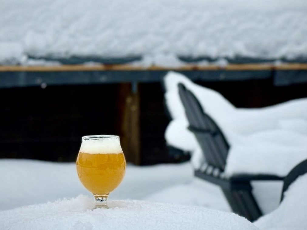 A tulip glass of beer sits chilled in a thick blanket of snow with snow-covered Adirondack chairs blurred in the background