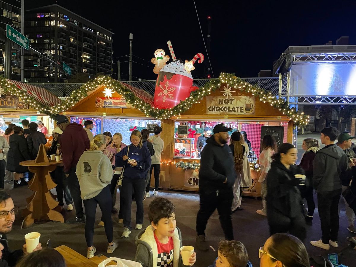 Crowds of holiday shoppers gather around festively decorated wooden vendor stalls at the Atlanta Christkindl Market at night, with a glowing hot chocolate booth and giant gingerbread man inflatable centerpiece lit up against the city skyline.