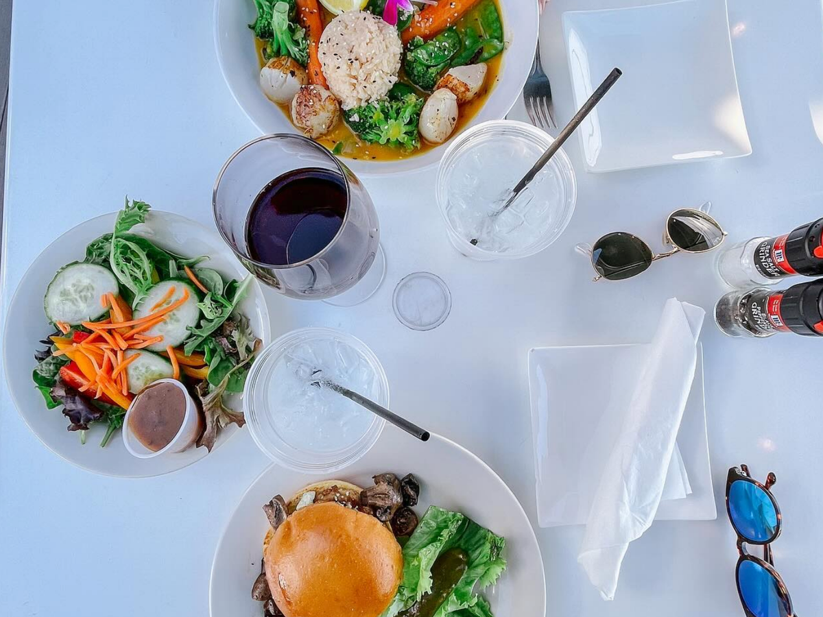 A vibrant outdoor lunch spread featuring a burger with lettuce and pickles, a colorful garden salad, and a vegetable and rice dish garnished with an edible flower, accompanied by drinks and sunglasses on a white table.