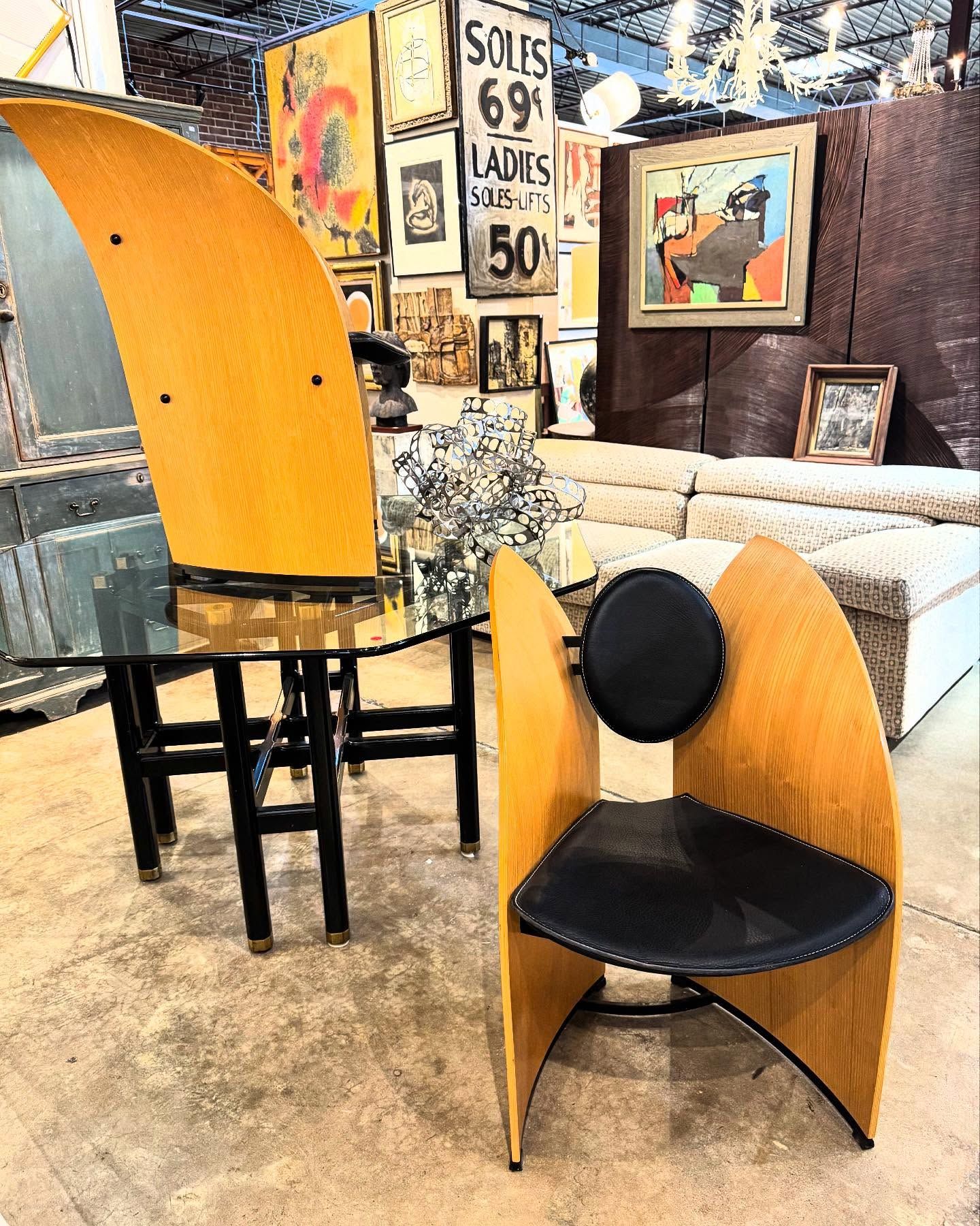 A  pair of sculptural mid-century modern chairs and a glass-topped table anchor a stylish vintage showroom at Trilogy Antiques & Design, surrounded by abstract artwork, a retro "Soles 69¢" sign, and a plush sectional sofa. Eclectic furnishings and eye-catching decor fill the warmly lit space.