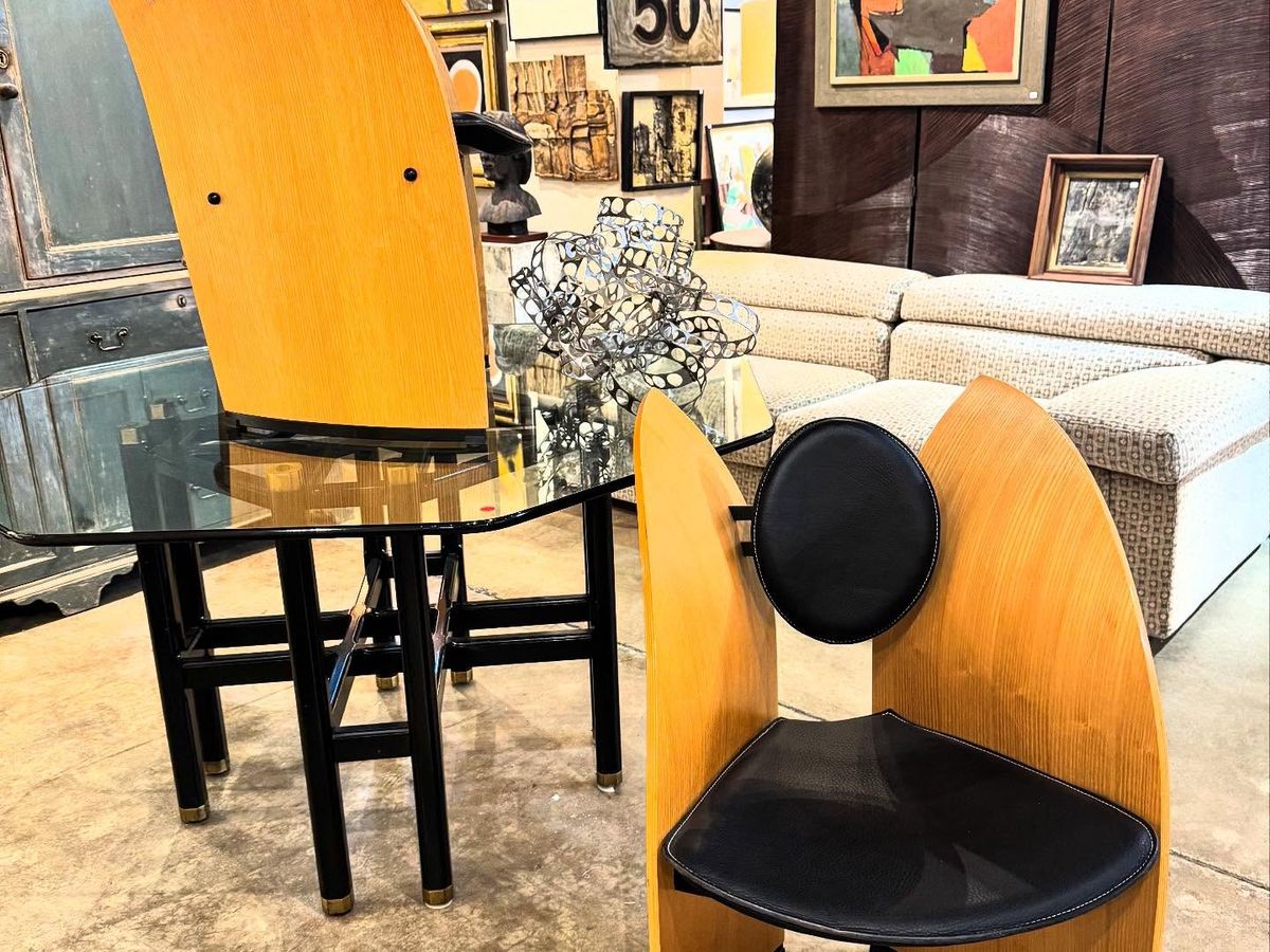 A pair of sculptural mid-century modern chairs and a glass-topped table anchor a stylish vintage showroom at Trilogy Antiques & Design, surrounded by abstract artwork, a retro "Soles 69¢" sign, and a plush sectional sofa. Eclectic furnishings and eye-catching decor fill the warmly lit space.