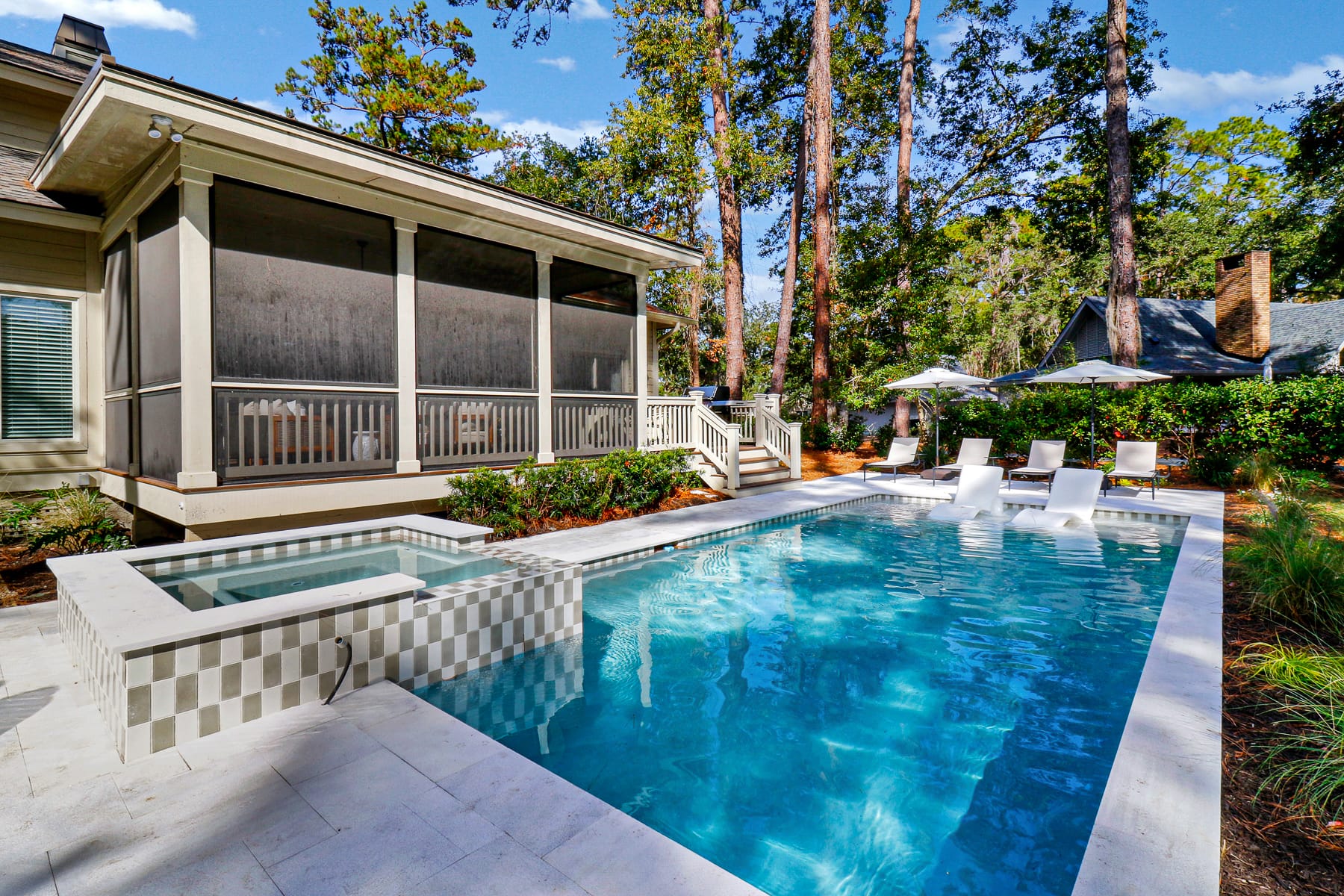 A newly renovated Sea Pines home features a modern open living room with large windows, a bright kitchen with a spacious island, and a sun-soaked pool and spa surrounded by a generous outdoor patio with wooden furniture.