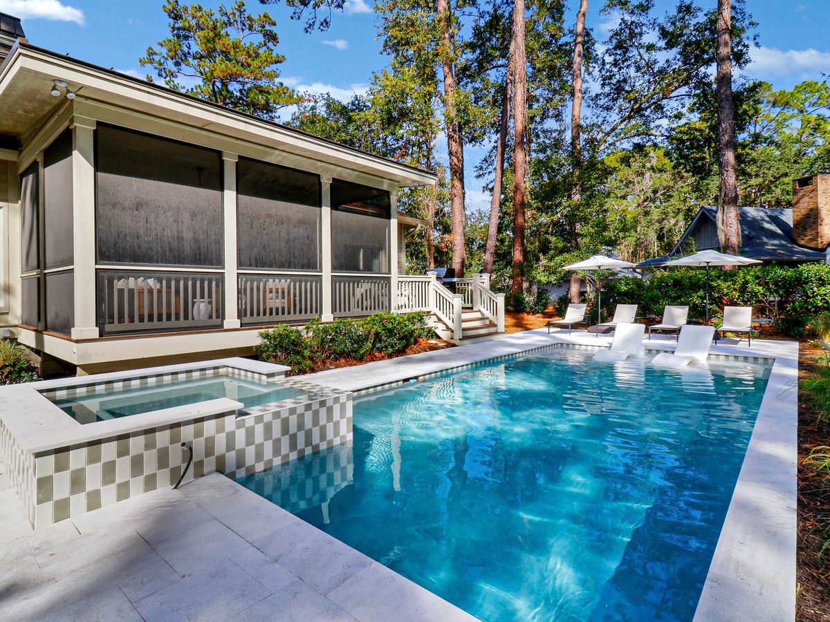 A newly renovated Sea Pines home features a modern open living room with large windows, a bright kitchen with a spacious island, and a sun-soaked pool and spa surrounded by a generous outdoor patio with wooden furniture.