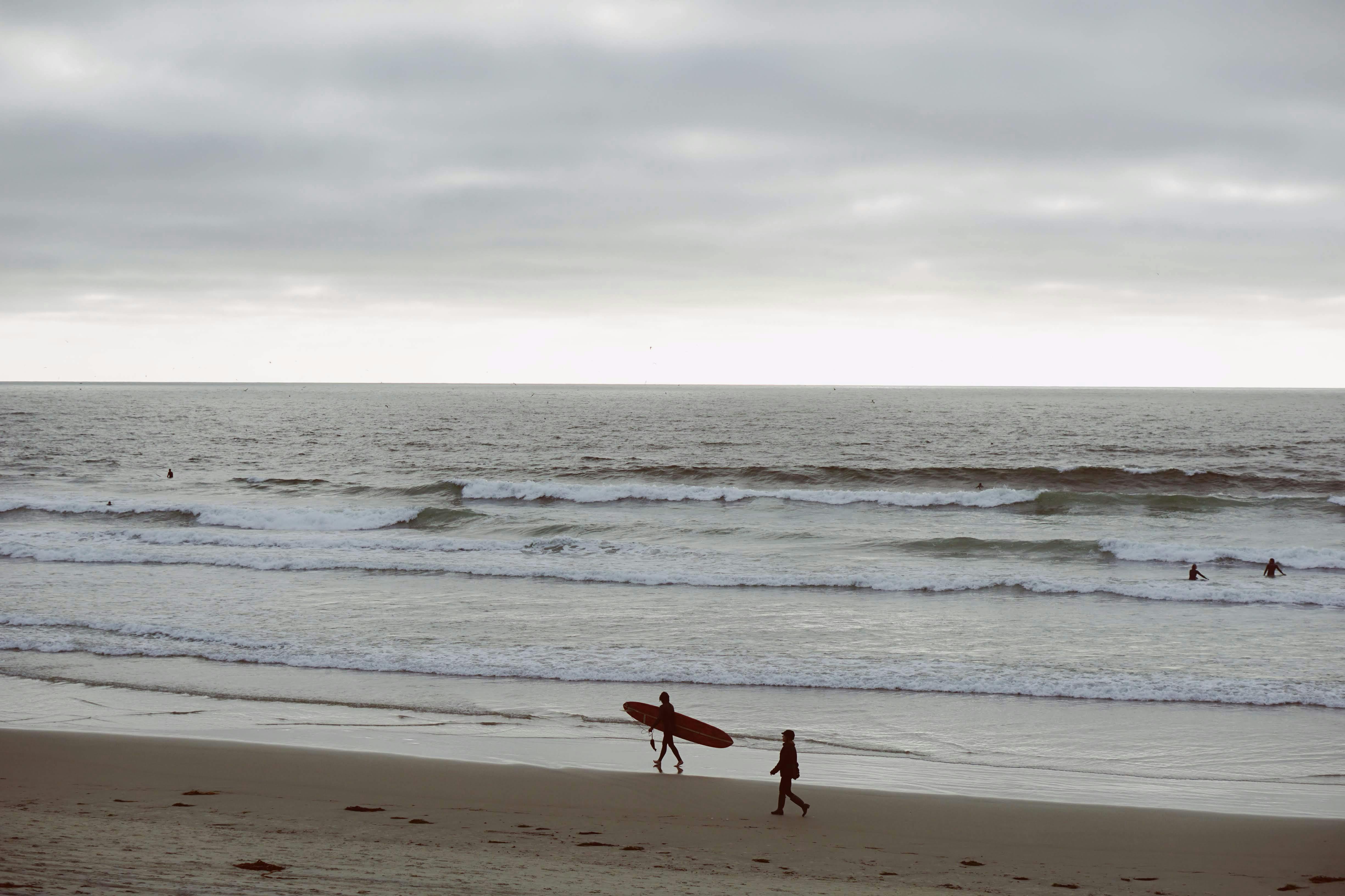 A surfer carries a longboard along the sandy shore while others ride waves in the background under a cloudy sky. This coastal scene captures the everyday mix of surfing and swimming activities at popular San Diego beaches.