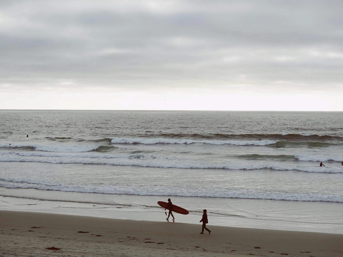 A surfer carries a longboard along the sandy shore while others ride waves in the background under a cloudy sky. This coastal scene captures the everyday mix of surfing and swimming activities at popular San Diego beaches.