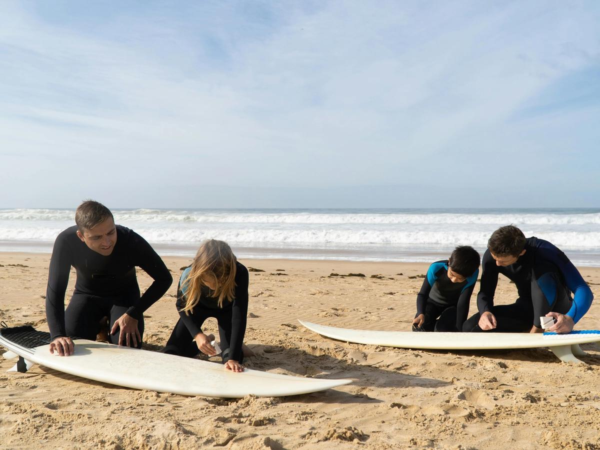 Four surfers kneel on the sand, waxing their boards as waves roll in behind them. The teamwork and ocean air show how staying active in nature lifts mood and confidence.