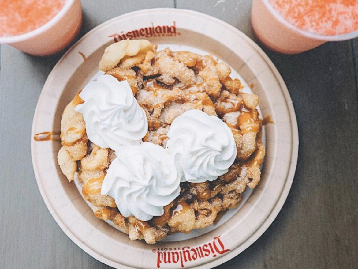Top-down view of a Disneyland funnel cake topped with whipped cream and caramel drizzle, served with two pink slushie drinks on a gray table.