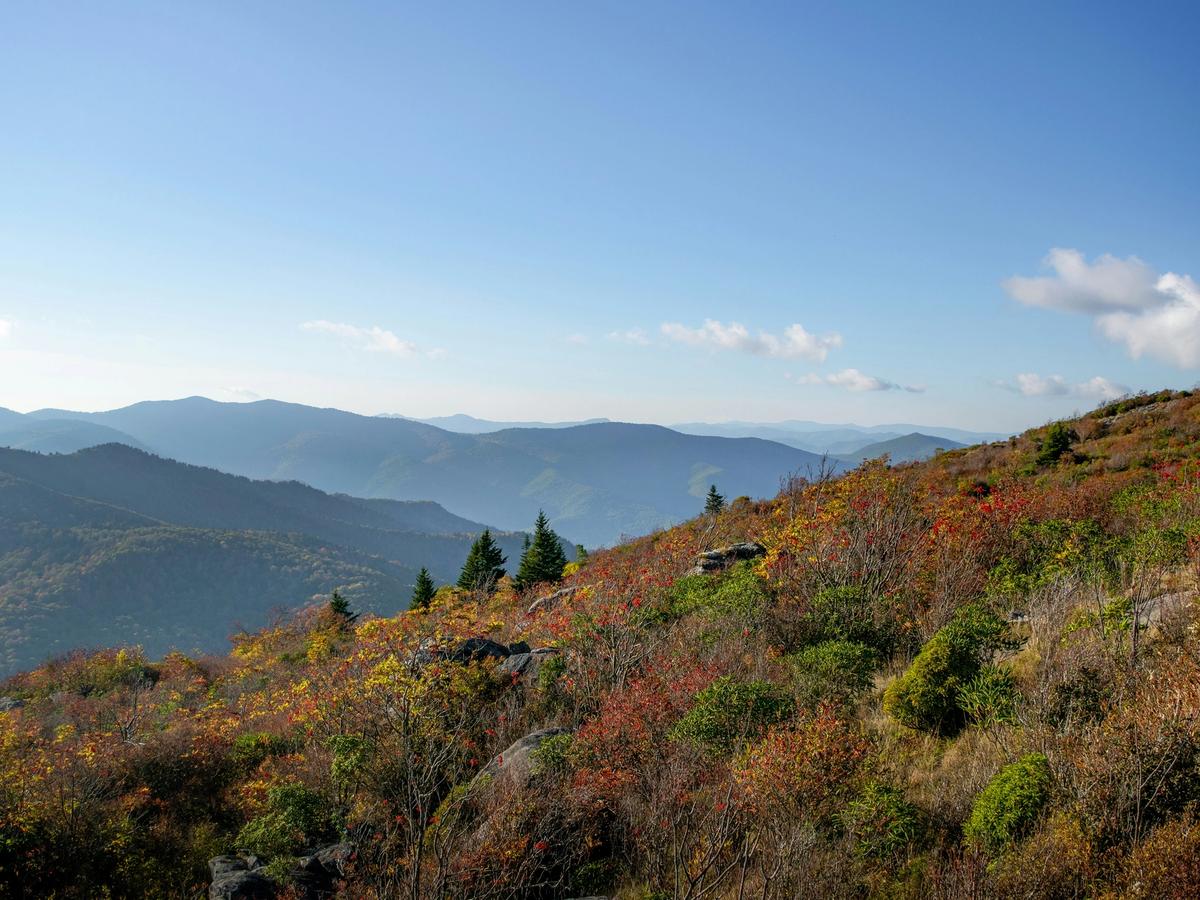 A scenic mountain landscape with rolling ridges fading into the distance, colorful autumn foliage on the hillside, and a bright blue sky with a few scattered clouds.