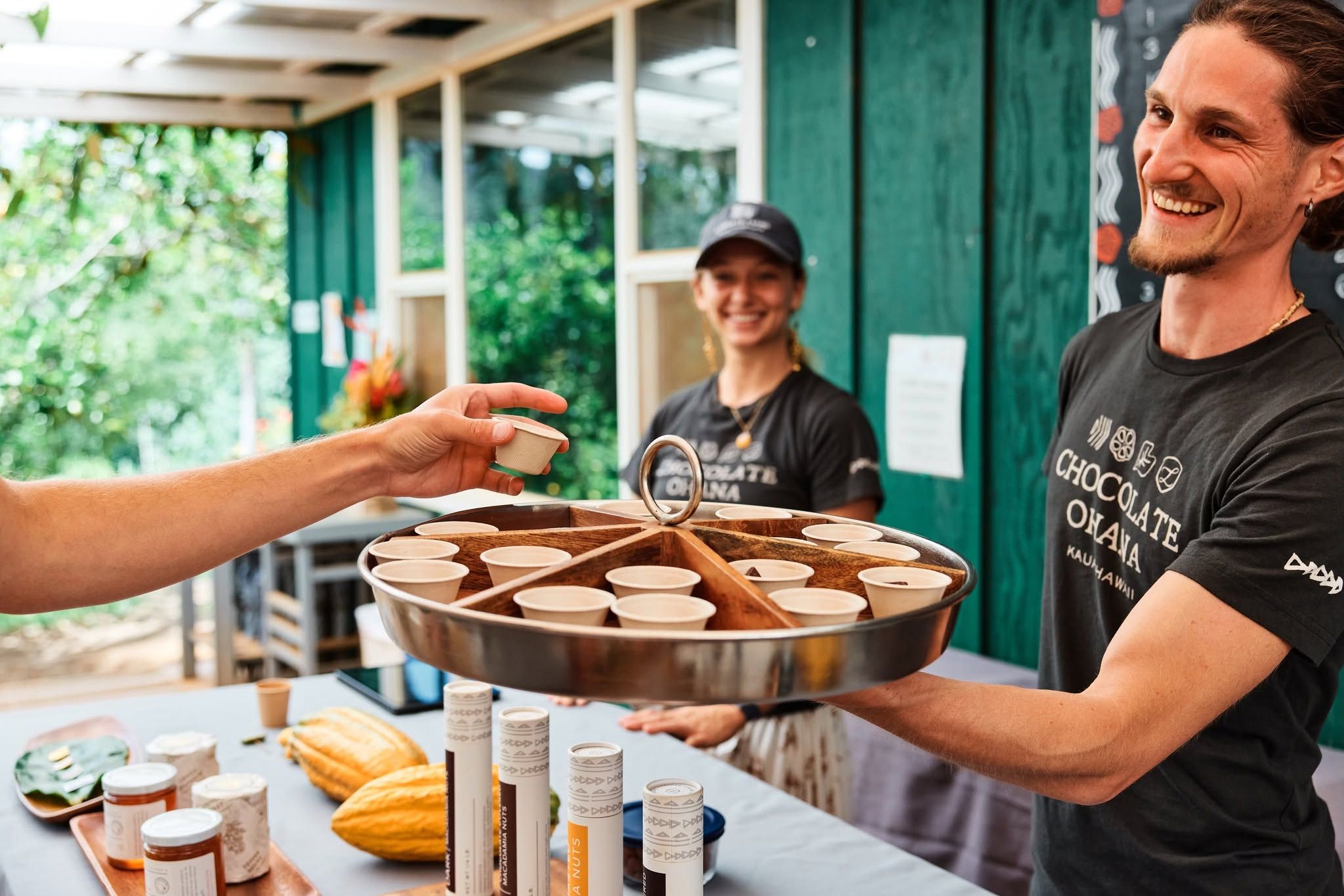 A smiling Lydgate Farms guide offers chocolate tasting samples to visitors on a farm tour in Kauai, Hawaii, with fresh cacao pods and Chocolate Ohana branded products displayed on the table.