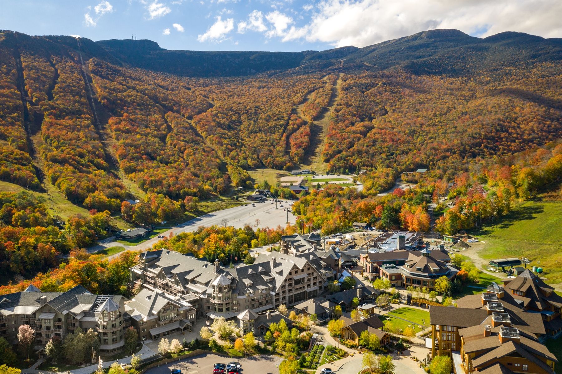 Aerial view of a picturesque mountain resort nestled at the base of tree-covered slopes bursting with fall foliage. 