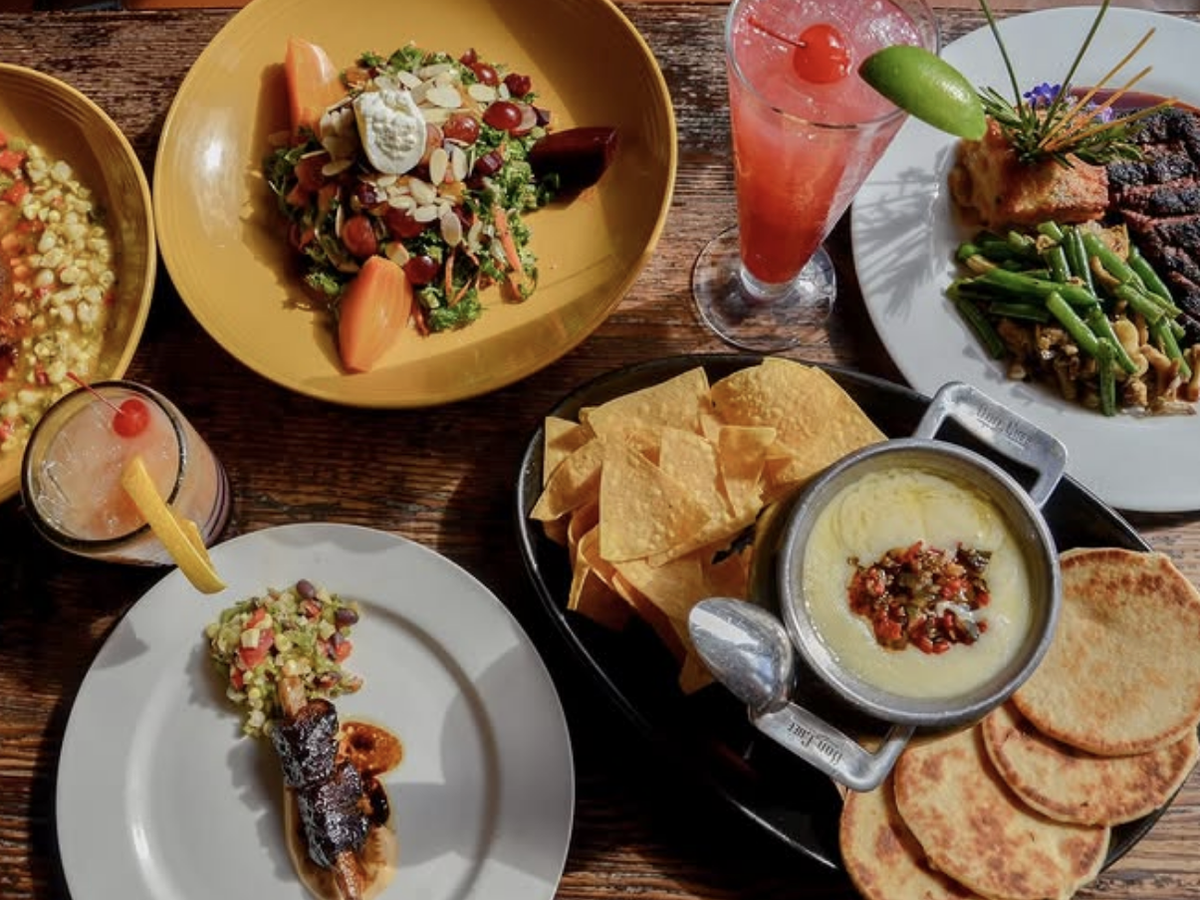 A colorful spread of gourmet dishes and drinks, including salad, grilled meats, queso with chips and flatbread, and vibrant cocktails on a rustic wooden table.