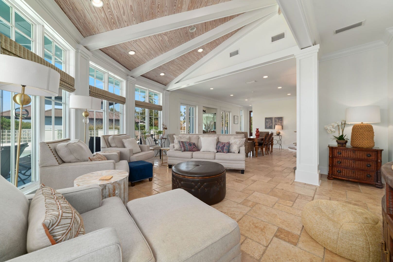 Elegant living space with vaulted wood ceilings, large windows, and neutral-toned seating. The open layout flows into a dining area, creating a bright and inviting setting.