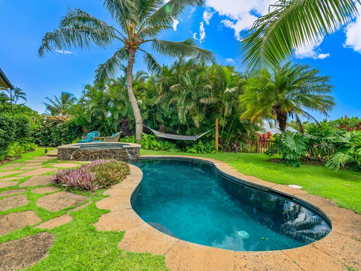 A tropical backyard oasis featuring a curved swimming pool, stone hot tub, hammock strung between palm trees, and lush greenery under a bright blue sky.