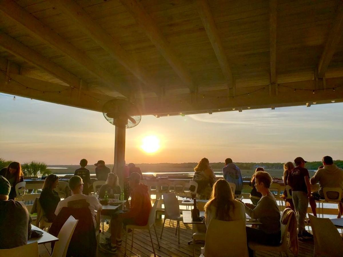 Guests sit and enjoy dinner at Jinks Creek Waterfront Grille as the sun sets over the water. The warm glow and open-air seating create a peaceful coastal dining experience.