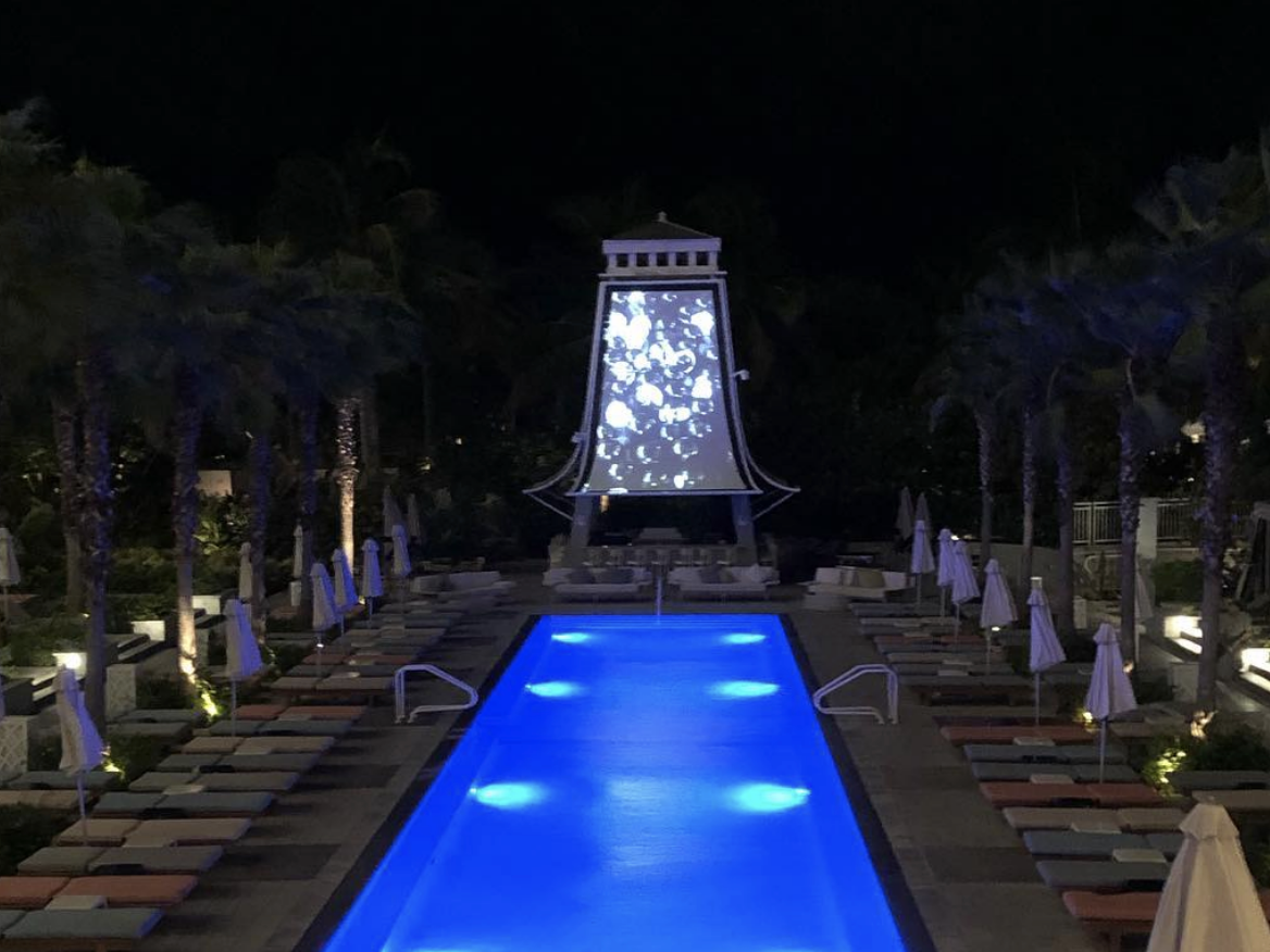 A sleek, illuminated pool glows blue at night, flanked by rows of lounge chairs and palm trees, with a striking screen structure at the end displaying a black-and-white projection.