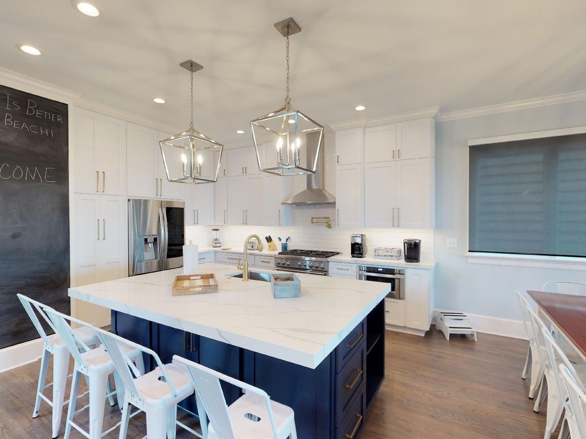 A spacious coastal kitchen with a large navy and white island, sleek white cabinetry, and stainless steel appliances. Modern pendant lights add elegance, while a chalkboard wall with 'Life is Better at the Beach!' welcomes guests. The open layout includes a wooden dining table with metal chairs, creating a stylish yet inviting space