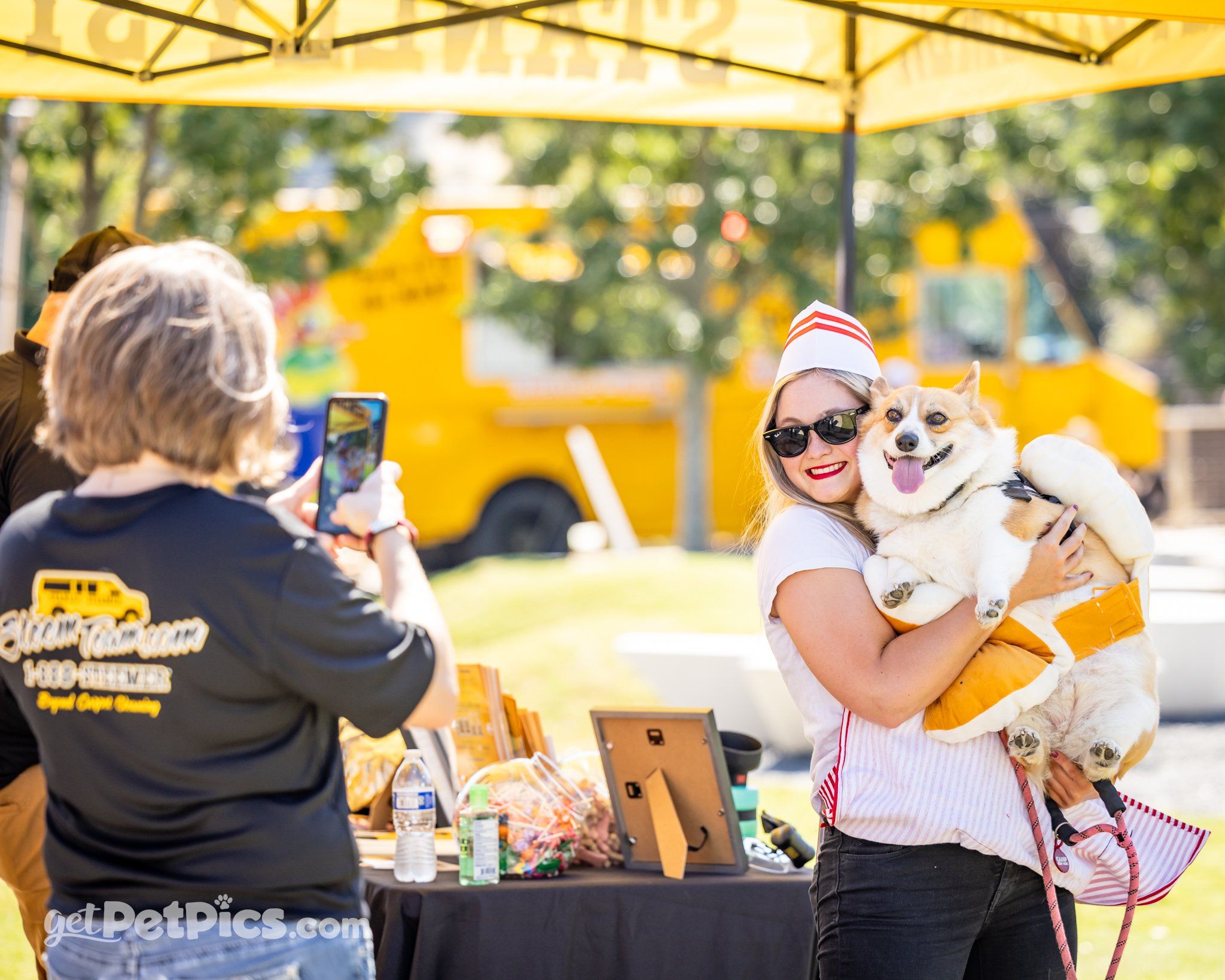 A woman dressed in a cute costume smiles as she holds her happy corgi in a fun Halloween outfit. Another person snaps a photo while a bright yellow food truck sets a cheerful backdrop. The moment feels playful and full of positive energy, perfect for a dog-friendly festival.