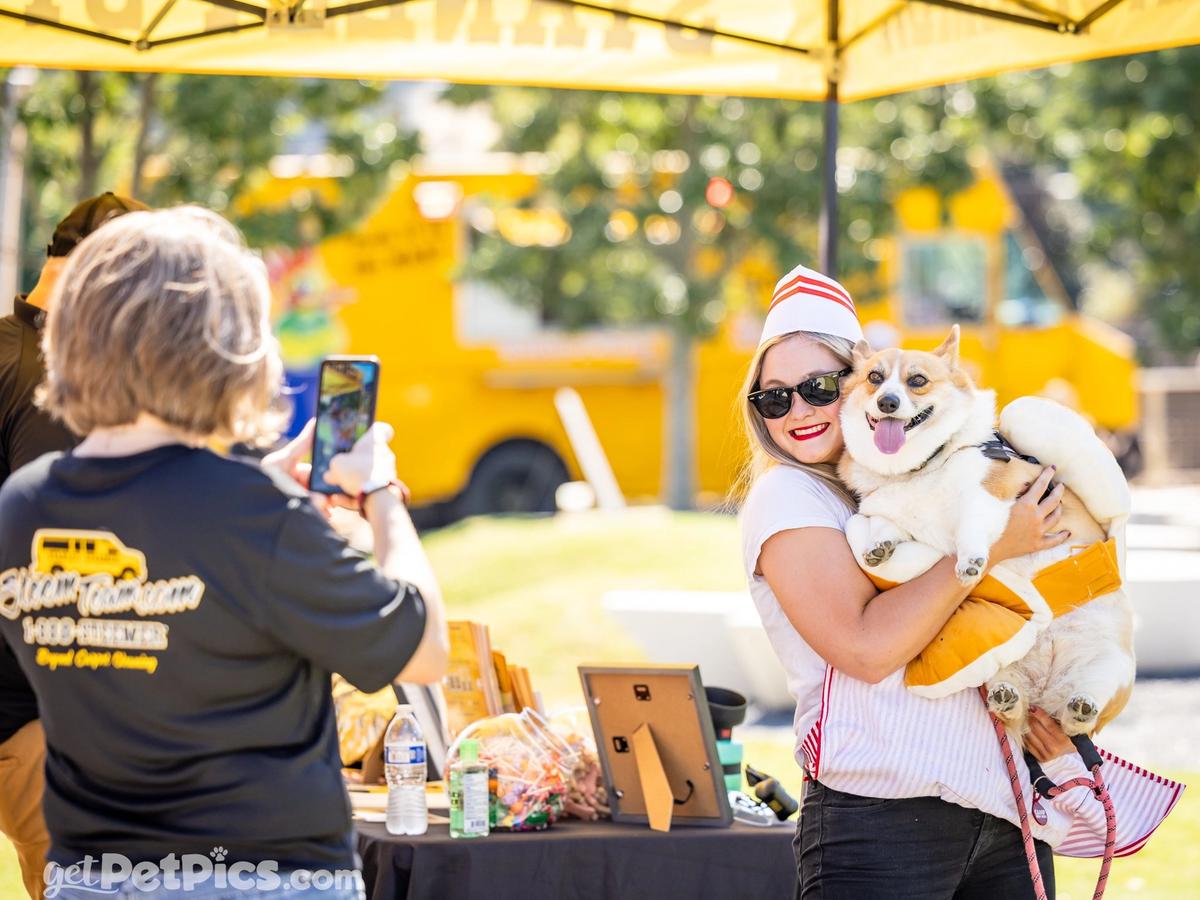 A woman dressed in a cute costume smiles as she holds her happy corgi in a fun Halloween outfit. Another person snaps a photo while a bright yellow food truck sets a cheerful backdrop. The moment feels playful and full of positive energy, perfect for a dog-friendly festival.