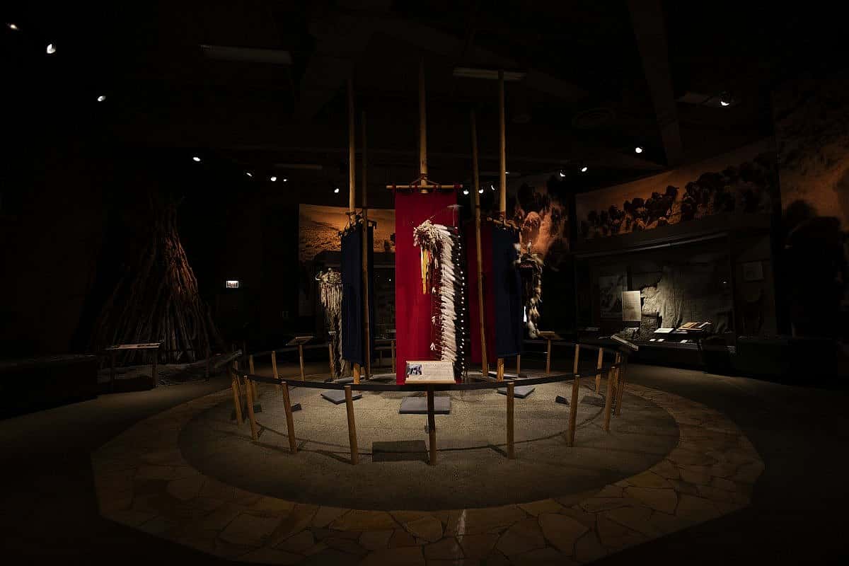 A dimly lit museum exhibit displays traditional Native American regalia, beadwork, and headdresses surrounded by storytelling panels and art. The scene honors the deep cultural history and craftsmanship of the Plains tribes.