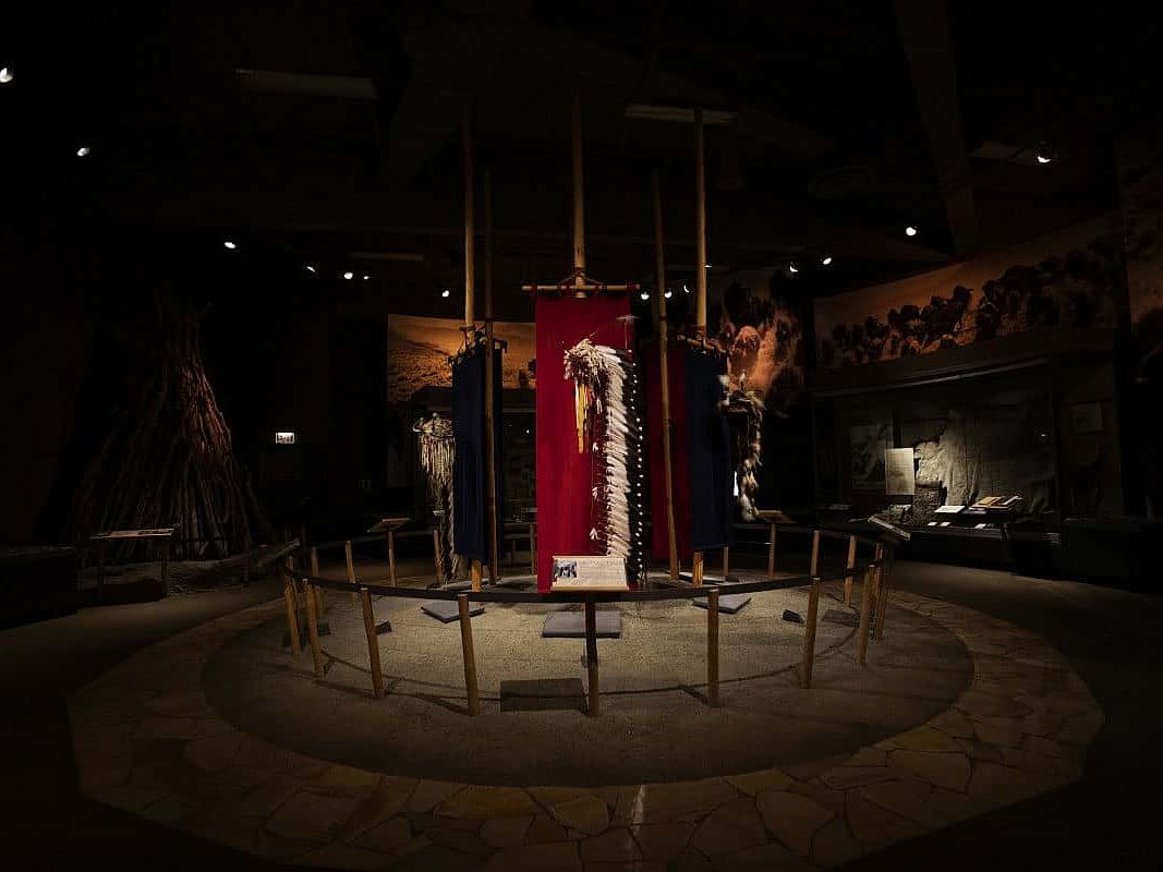 A dimly lit museum exhibit displays traditional Native American regalia, beadwork, and headdresses surrounded by storytelling panels and art. The scene honors the deep cultural history and craftsmanship of the Plains tribes.