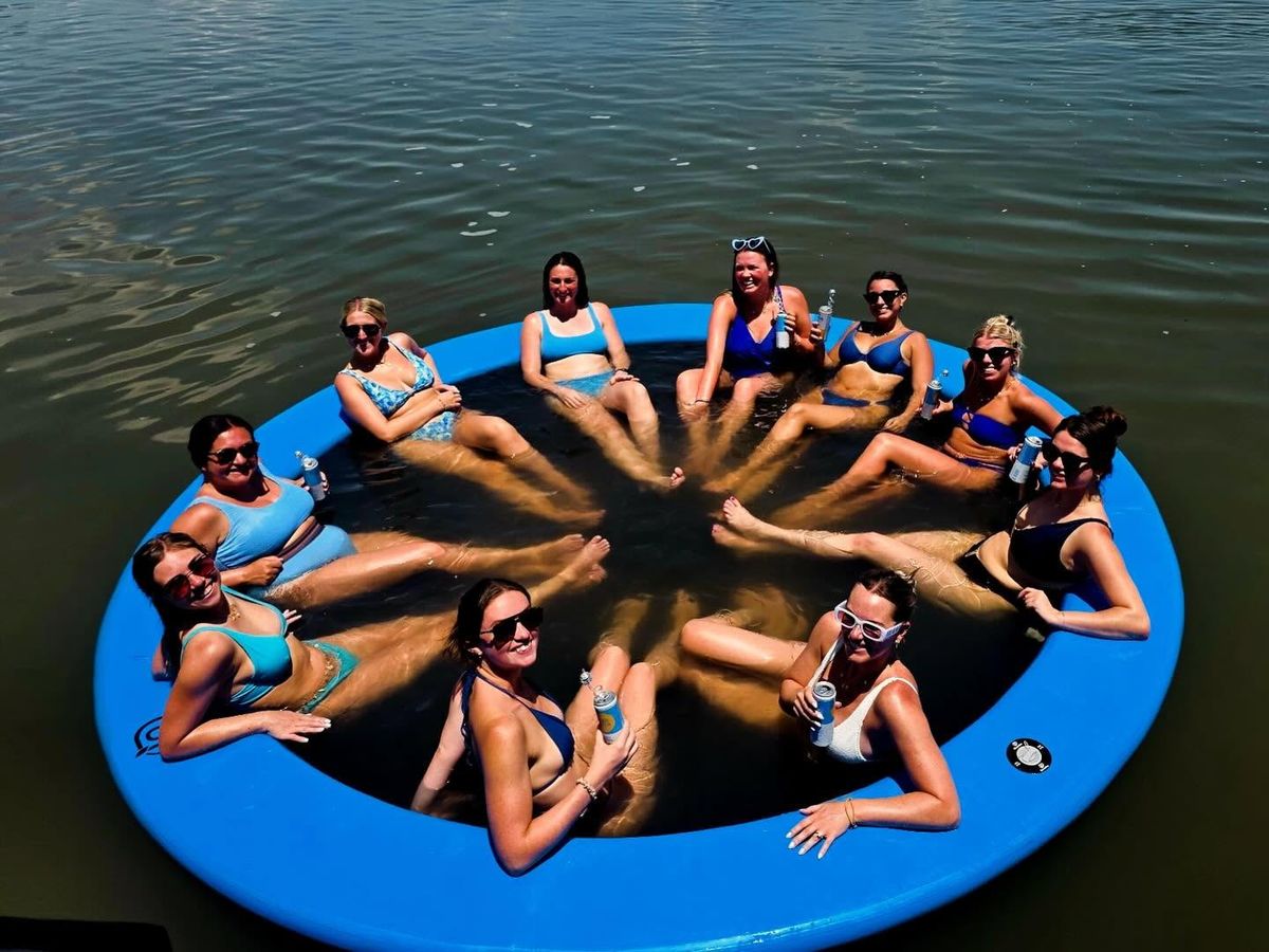 group of women in swimsuits and sunglasses relax together in a large circular blue floating lounger on calm water, their feet meeting at the center as they hold drinks and smile. Sunlight sparkles across the surrounding water on a bright summer day.