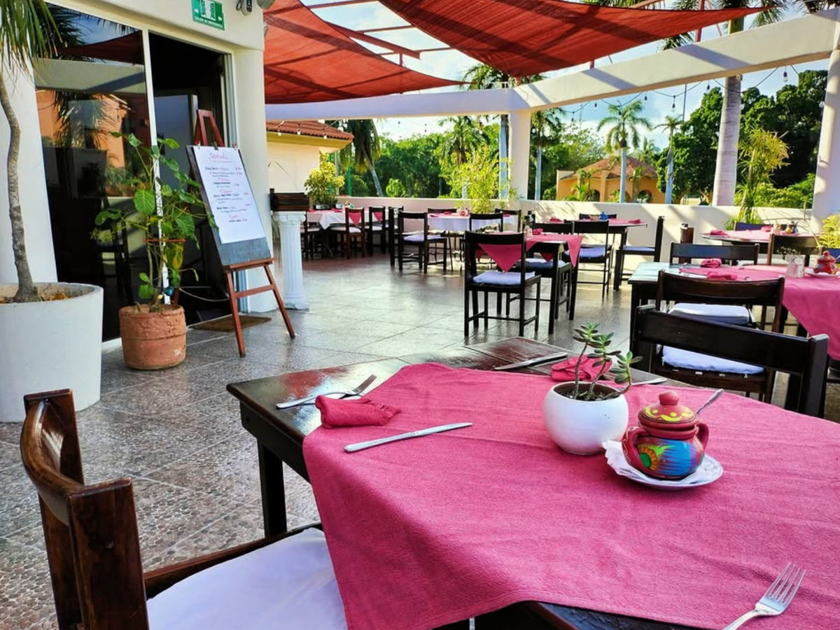 An inviting open-air restaurant patio with tables covered in pink tablecloths, set for dining with utensils, potted plants, and a colorful ceramic condiment jar, surrounded by tropical greenery and shaded by red canopies.
