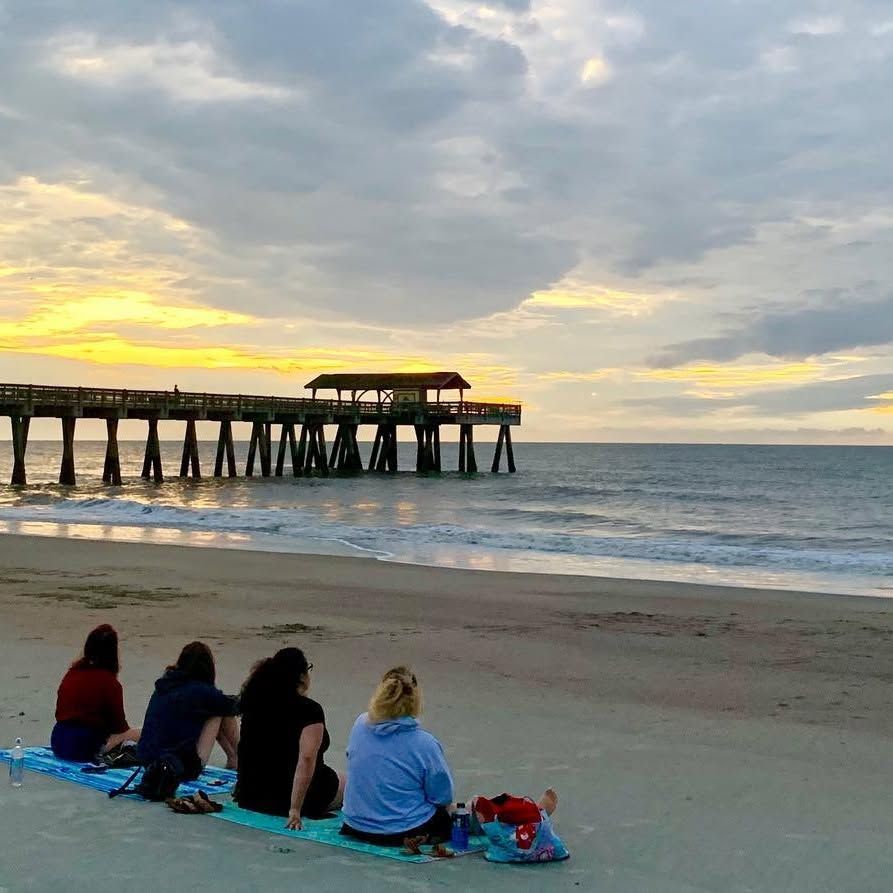 Four people sit together on a beach towel watching a golden sunrise over the ocean beside the Tybee Island Pier. Soft clouds glow orange and gray above the calm water as gentle waves roll toward the shore. 