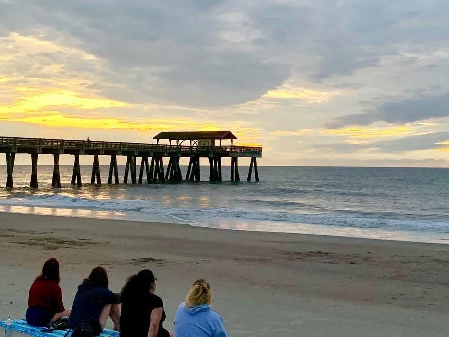 Four people sit together on a beach towel watching a golden sunrise over the ocean beside the Tybee Island Pier. Soft clouds glow orange and gray above the calm water as gentle waves roll toward the shore.