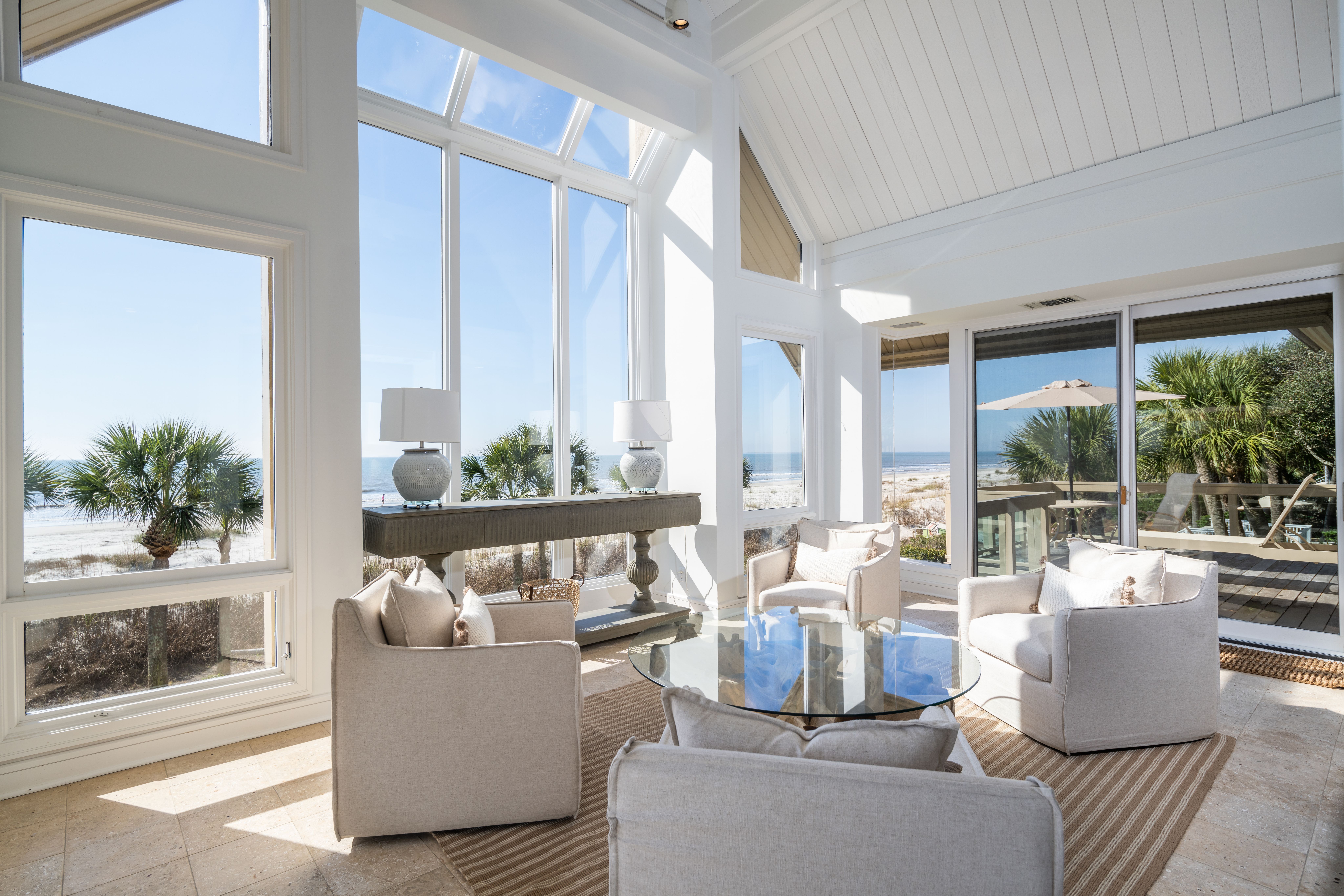 A bright coastal living room with tall windows and a vaulted ceiling offers panoramic views of the beach and palm trees, featuring neutral-toned armchairs, a glass coffee table, and soft natural light.