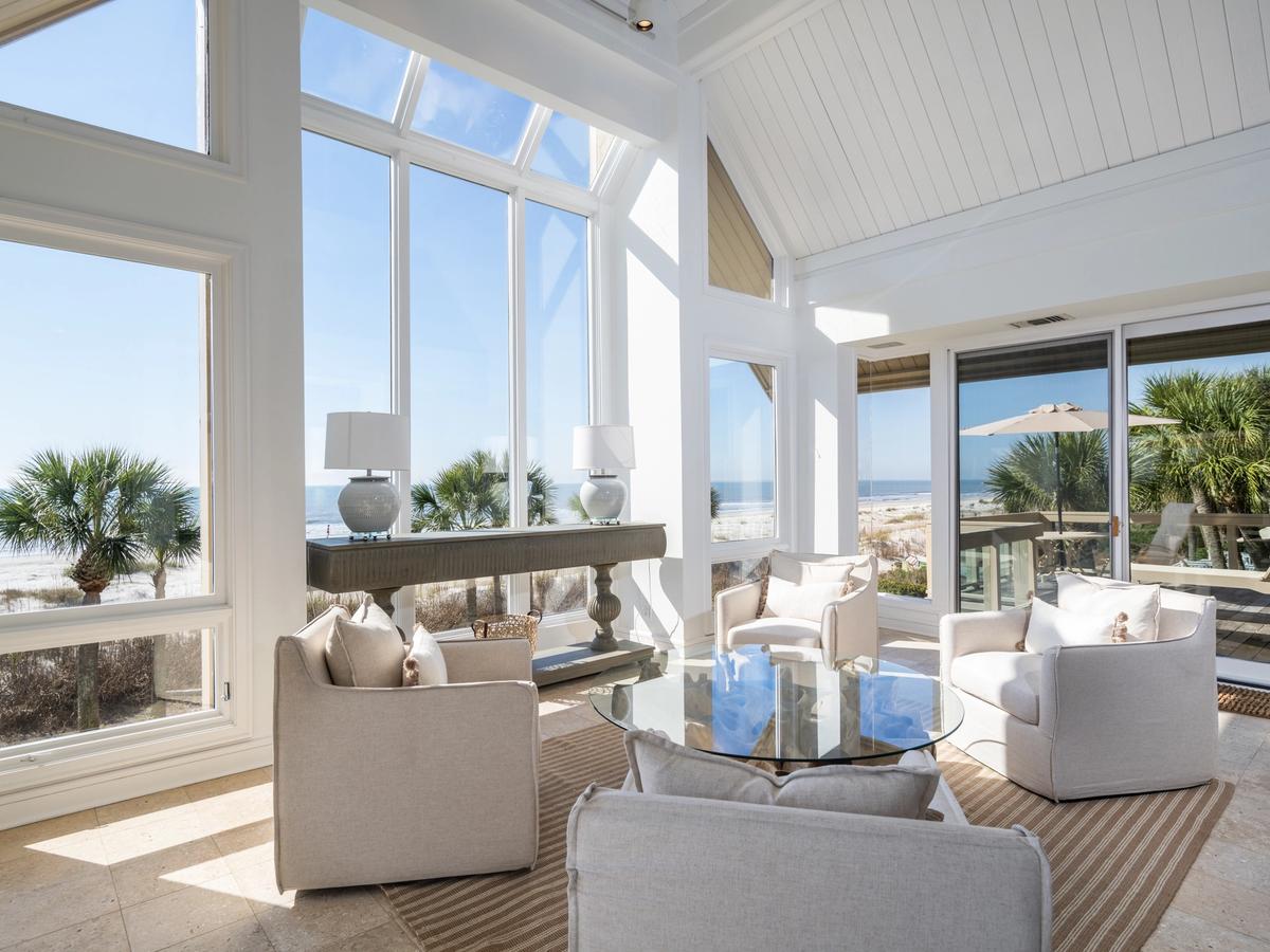 A bright coastal living room with tall windows and a vaulted ceiling offers panoramic views of the beach and palm trees, featuring neutral-toned armchairs, a glass coffee table, and soft natural light.