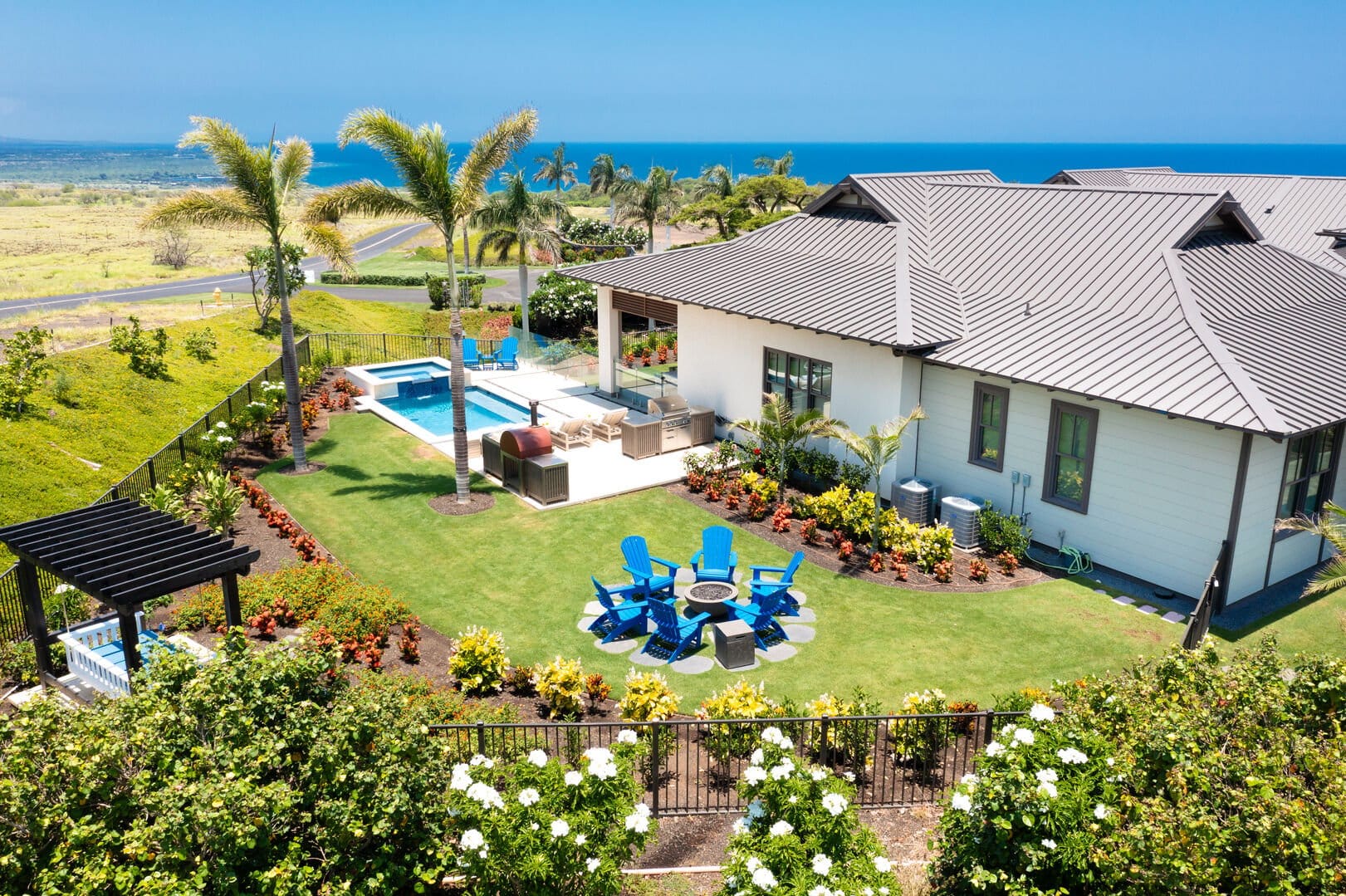 Blue Serenity on Hawaii’s Big Island has a pool and a green lawn overlooking the ocean. Blue chairs surround a fire pit, with palm trees and flowering plants lining the yard.