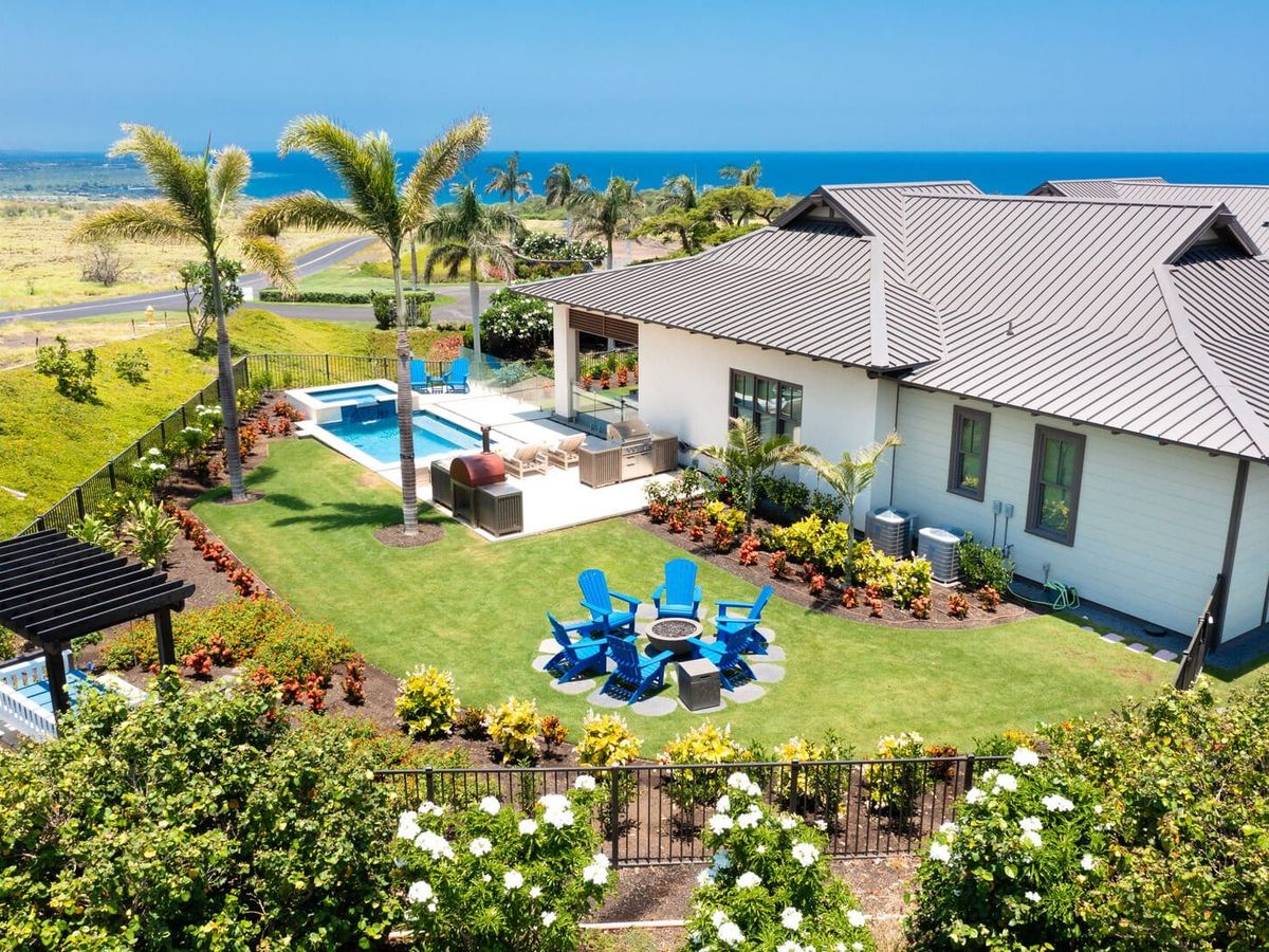 Blue Serenity on Hawaii’s Big Island has a pool and a green lawn overlooking the ocean. Blue chairs surround a fire pit, with palm trees and flowering plants lining the yard.