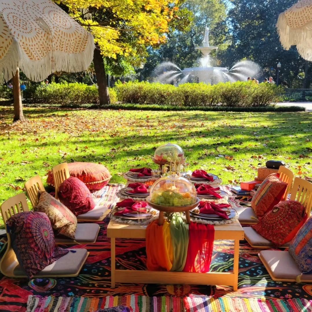 even women dressed in stylish spring outfits stand together and smile behind a beautifully styled outdoor picnic table set with fresh sandwiches, floral centerpieces, plush pillows, and elegant place settings. Spanish moss drapes from the trees of a lush green park, with a mounted horse visible in the background.