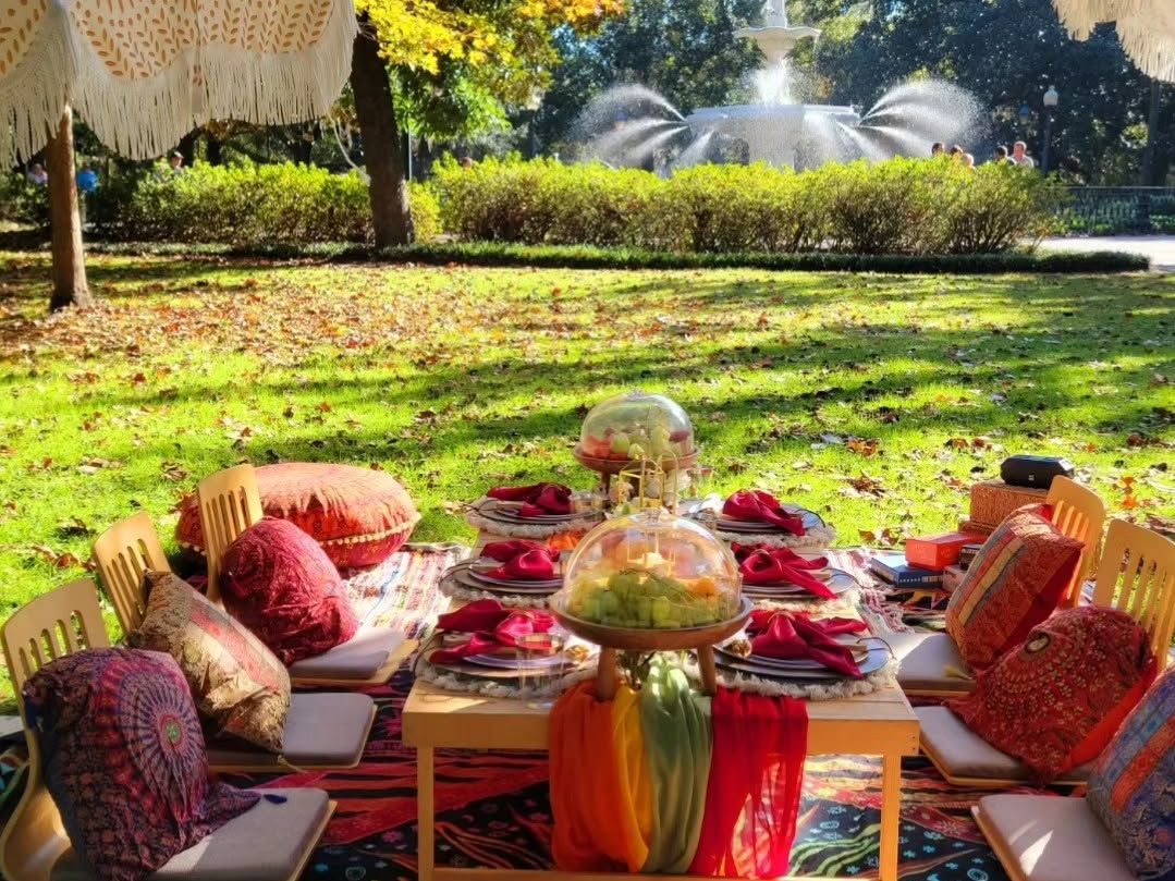 even women dressed in stylish spring outfits stand together and smile behind a beautifully styled outdoor picnic table set with fresh sandwiches, floral centerpieces, plush pillows, and elegant place settings. Spanish moss drapes from the trees of a lush green park, with a mounted horse visible in the background.