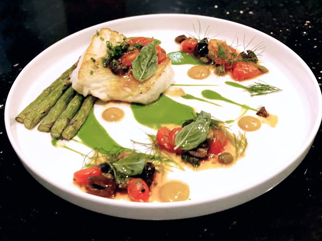 A beautifully plated cod fillet with grilled asparagus and bright herb sauces. This elegant dish is finished with olives, cherry tomatoes, and fresh basil for a balanced bite.