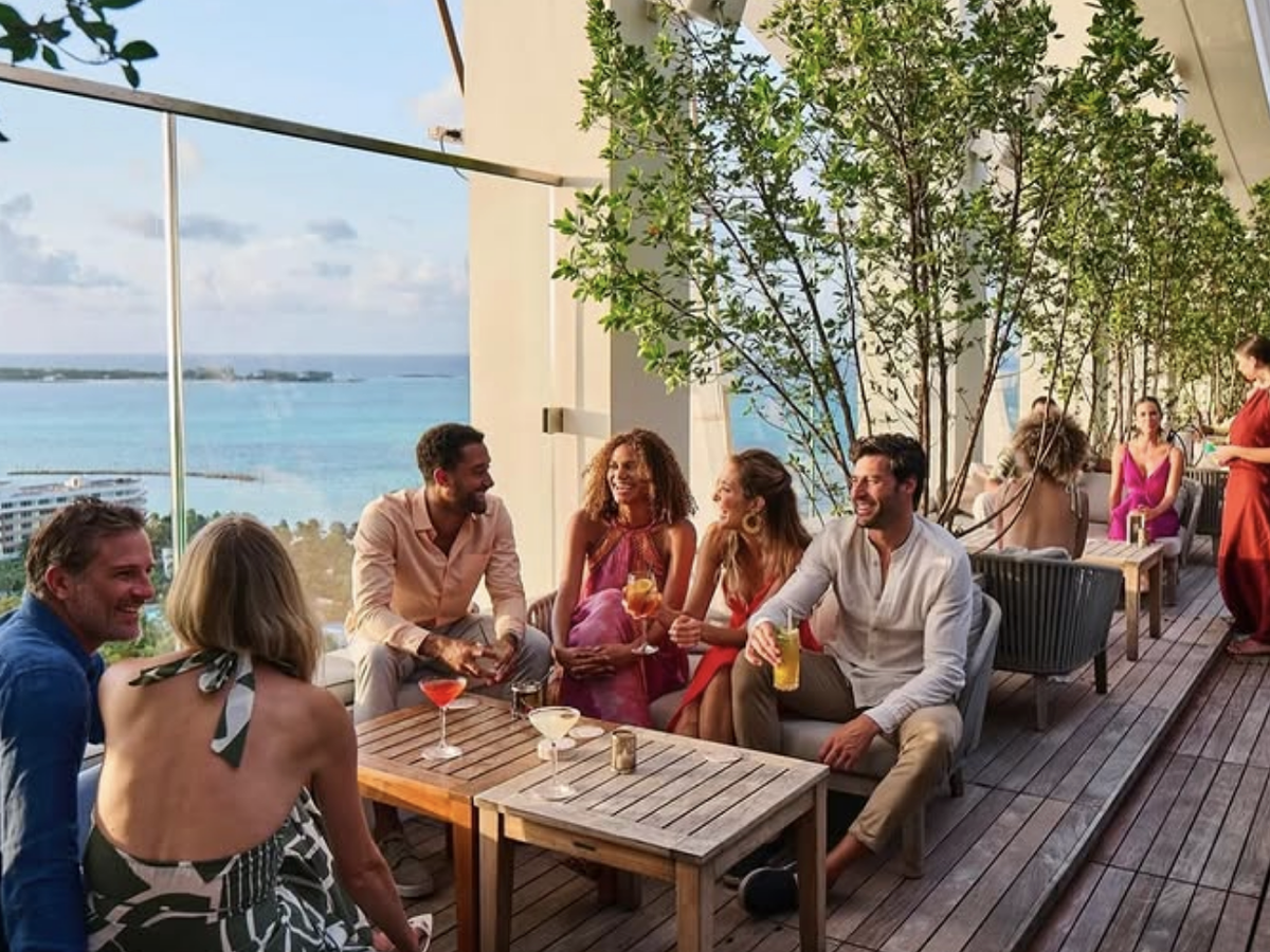 A group of friends relaxes and laughs while enjoying cocktails on a stylish rooftop lounge with ocean views, lush greenery, and warm evening light.