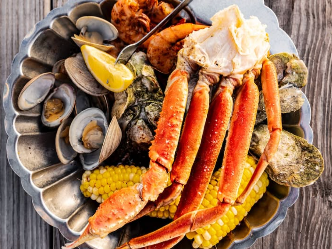 A seafood platter featuring crab legs, shrimp, clams, oysters, corn on the cob, and a lemon wedge, served on a metal dish over a rustic wooden table.