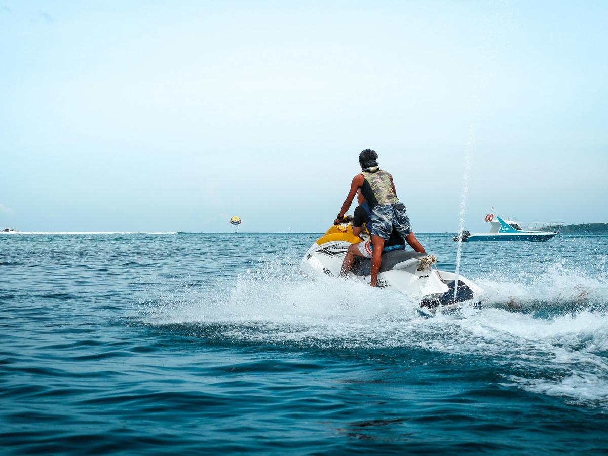 Guy standing up on back of jetski while front person drives it and boat in the background