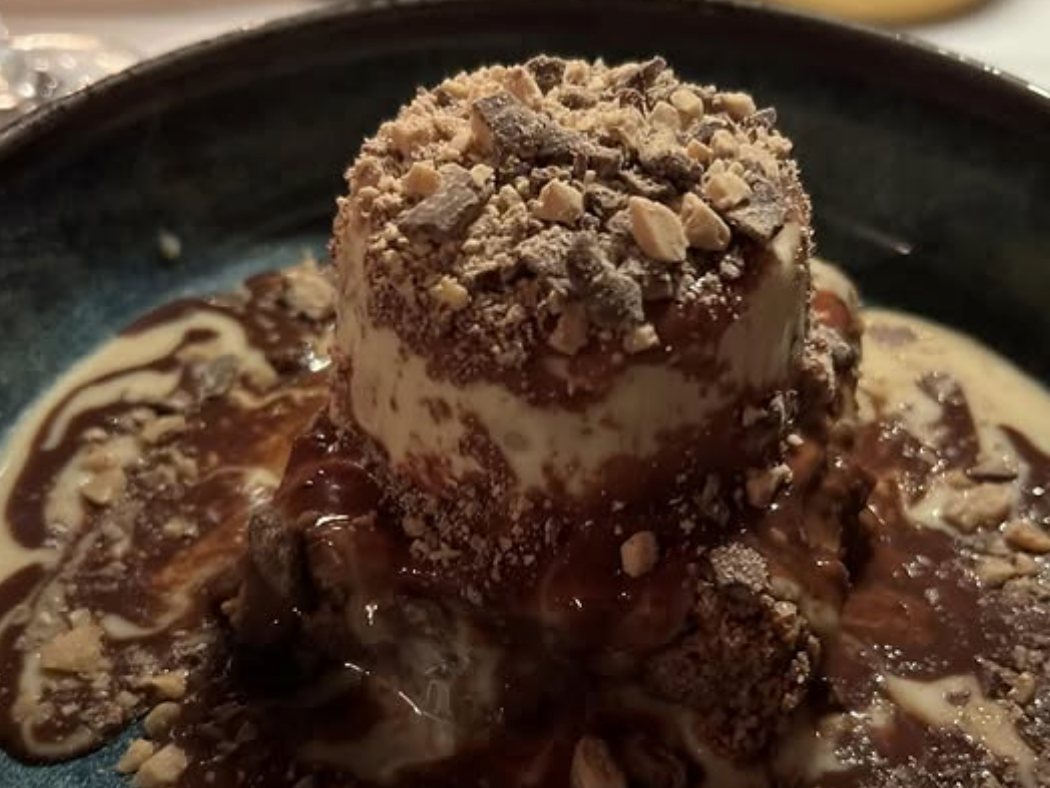 A decadent dessert of vanilla ice cream coated in chocolate sauce and crushed toffee bits sits melting in a dark ceramic bowl.